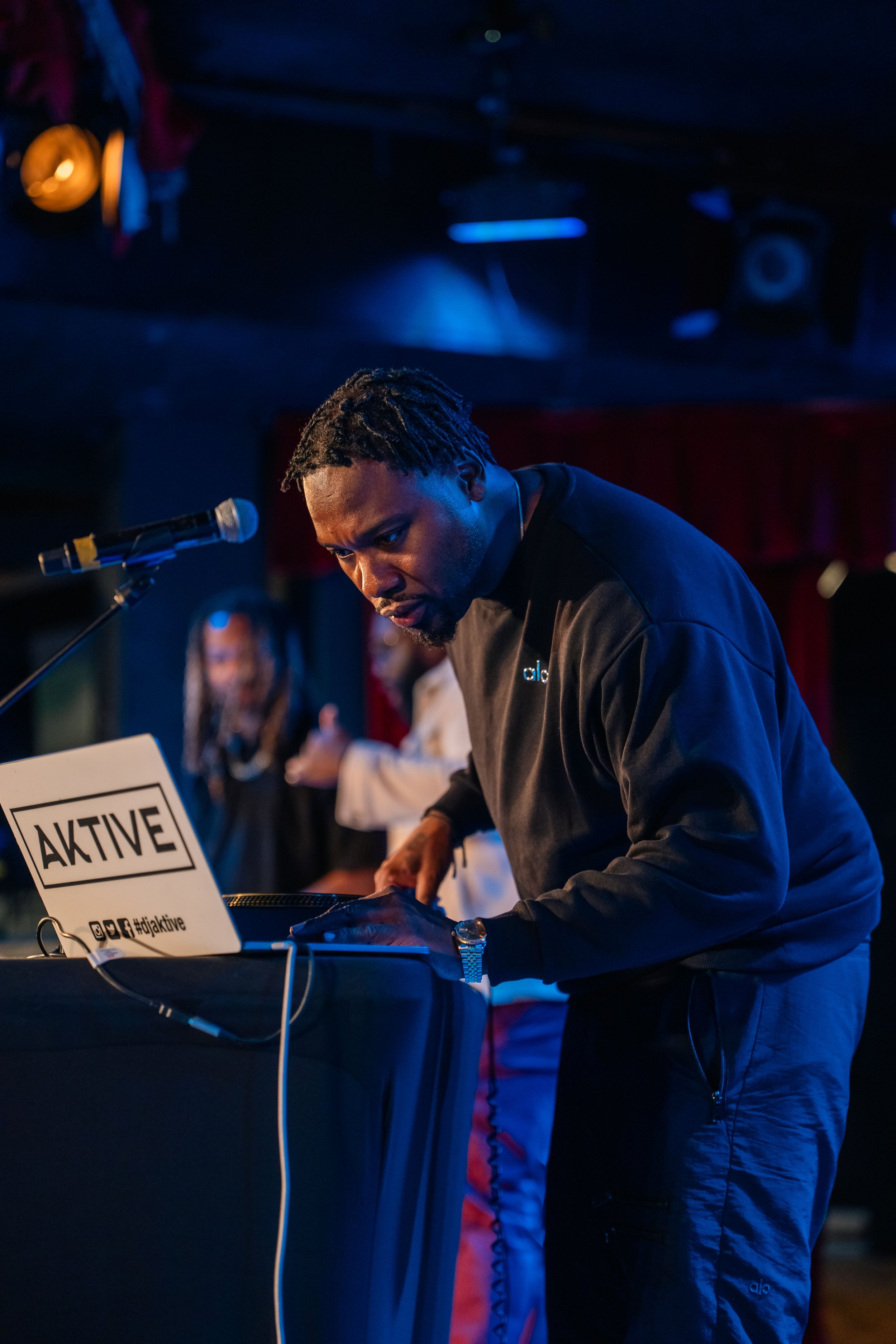 A man with dark skin and dreadlocks wearing a black jacket leaning over a table with a microphone in front of him. There is a sign on the table that says 'AKTIVE' and has social media icons. In the blurred background, a woman and a man are visible. T