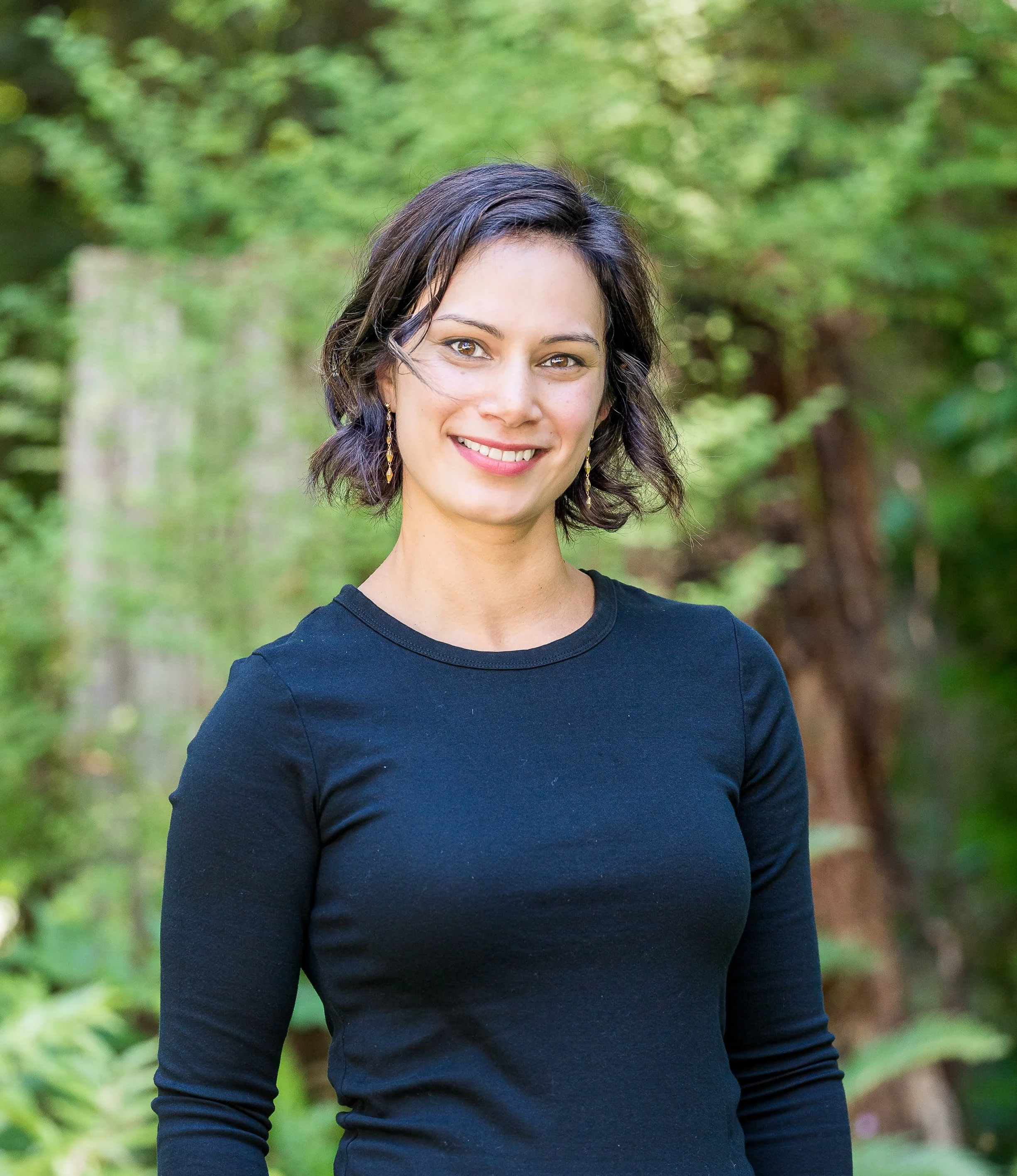 A smiling woman with short dark hair, wearing a black long-sleeve top and gold earrings, standing outdoors in a wooded area with green trees in the background.