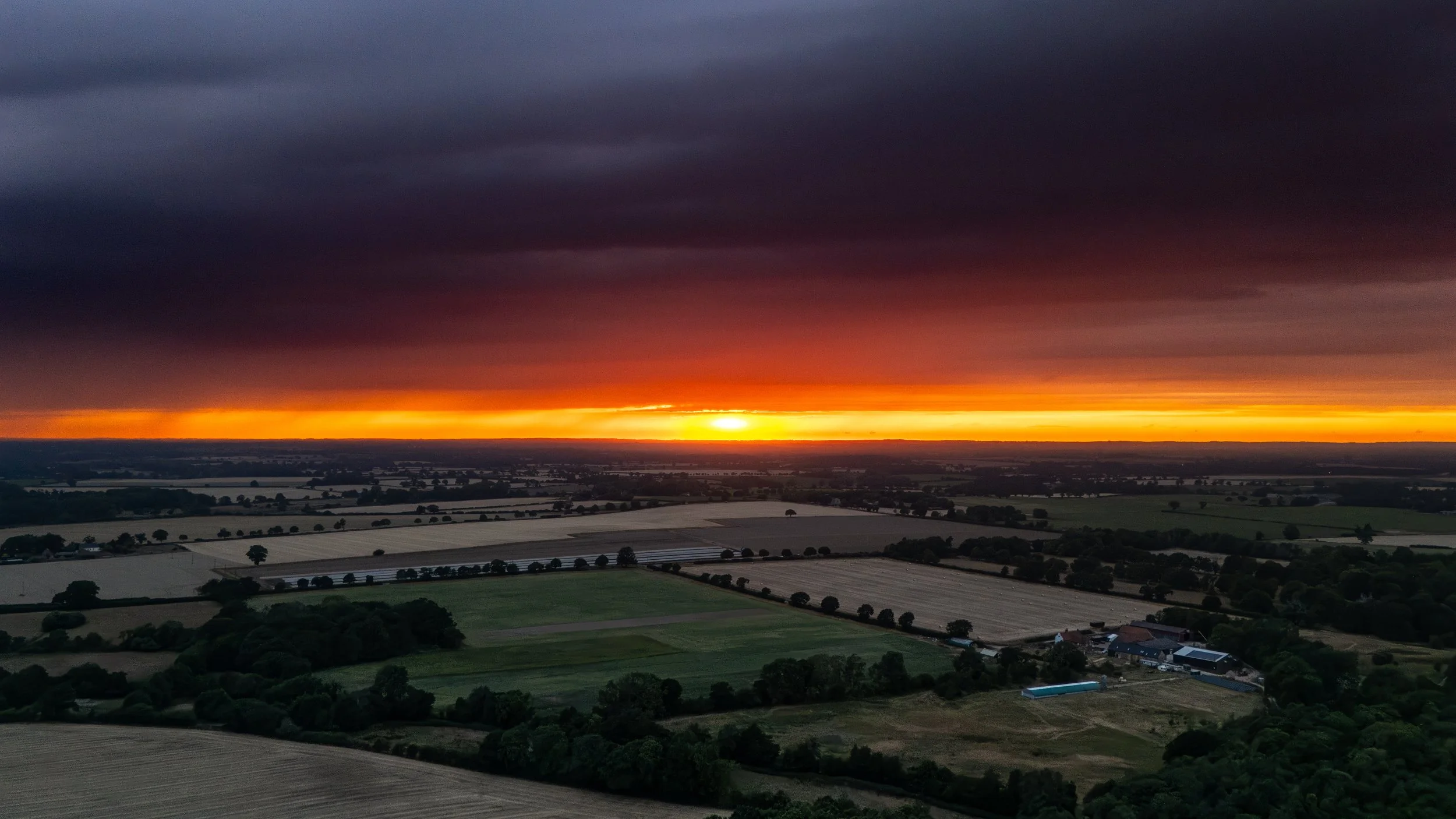 A view of a sunset over farmland with dark storm clouds overhead, illuminated by the orange and yellow glow of the setting sun, with fields, trees, and farm buildings below.