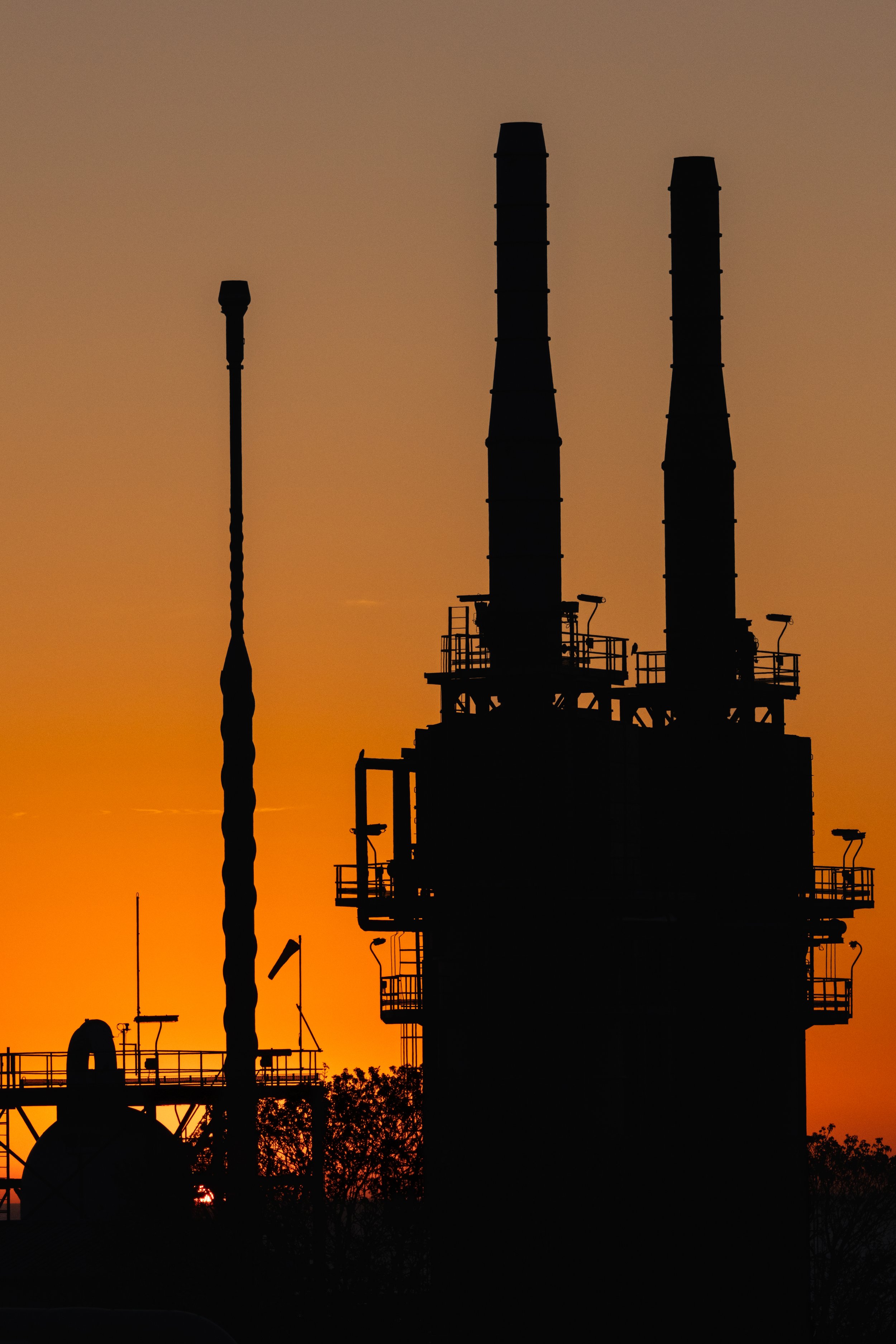 Silhouette of an industrial processing facility with tall stacks and equipment against an orange sunset sky.