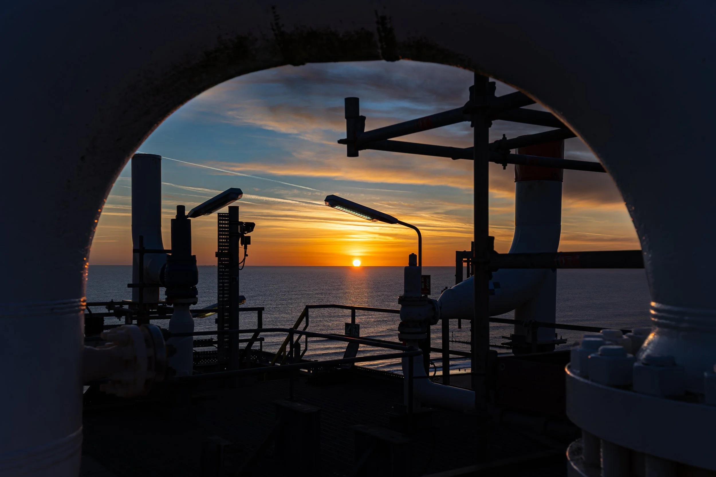 Sunset view over the ocean framed by industrial pipes and structures on a gas platform.