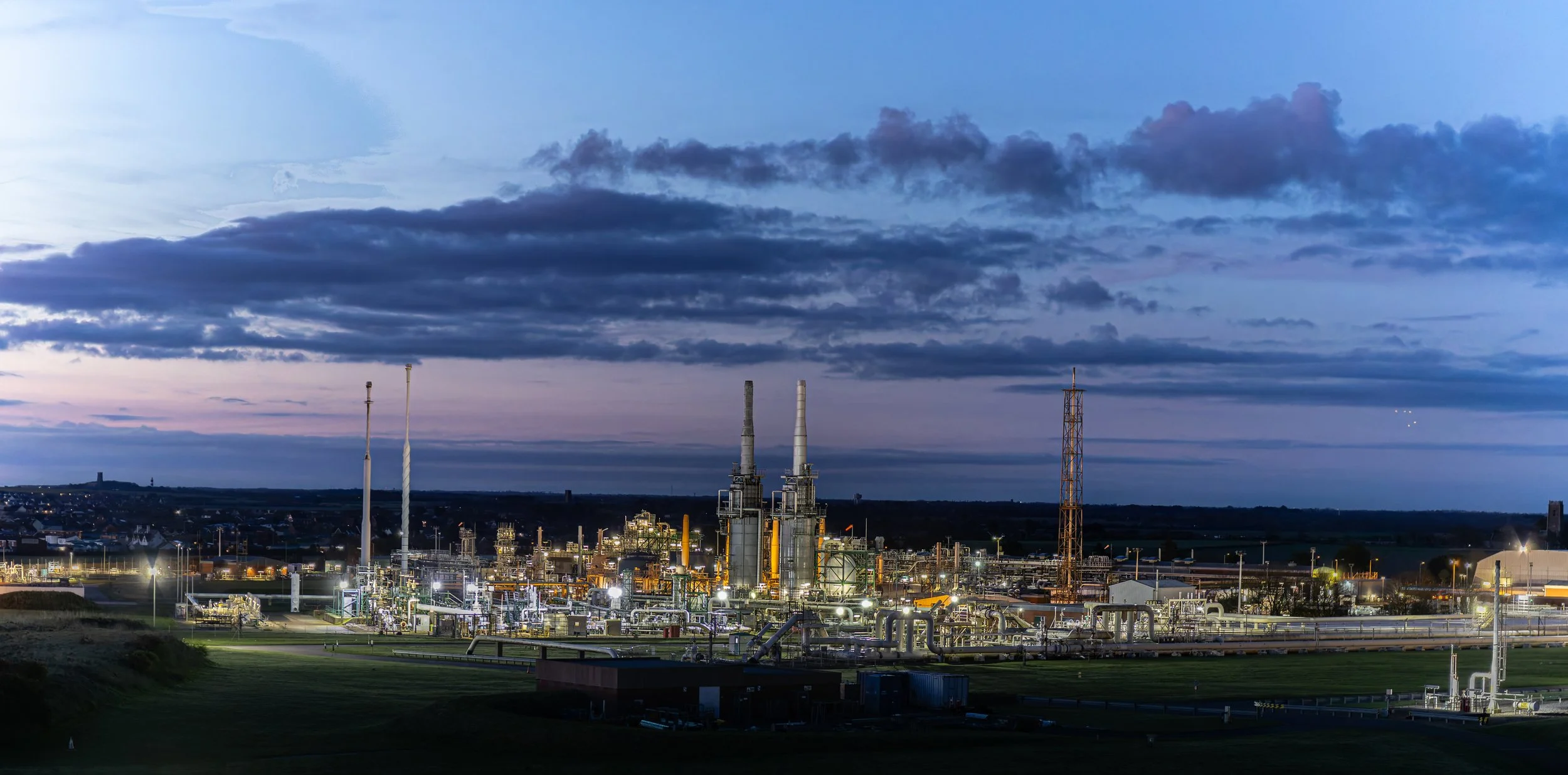 An industrial processing facility with multiple tall towers and pipes, illuminated against a cloudy evening sky.
