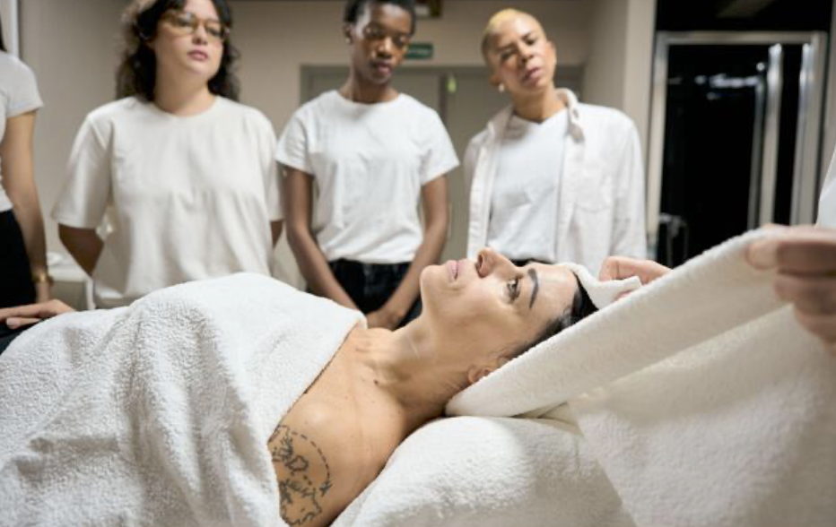 A woman lying in an Esthetician bed with three women standing around her, discussing treatment.