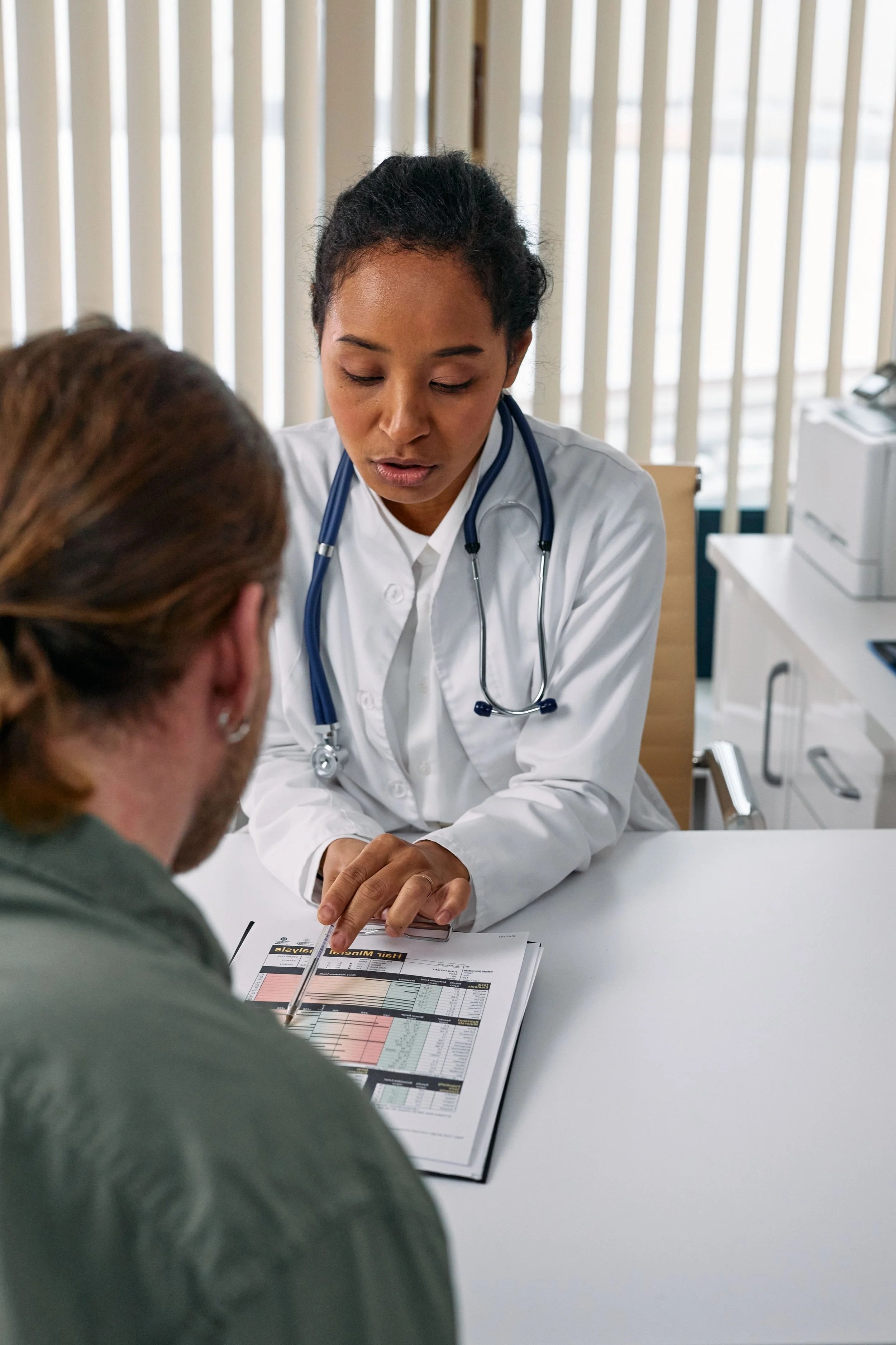 Medical professional in white coat and stethoscope consulting with a patient at a desk, reviewing medical charts or documents in a clinical setting.