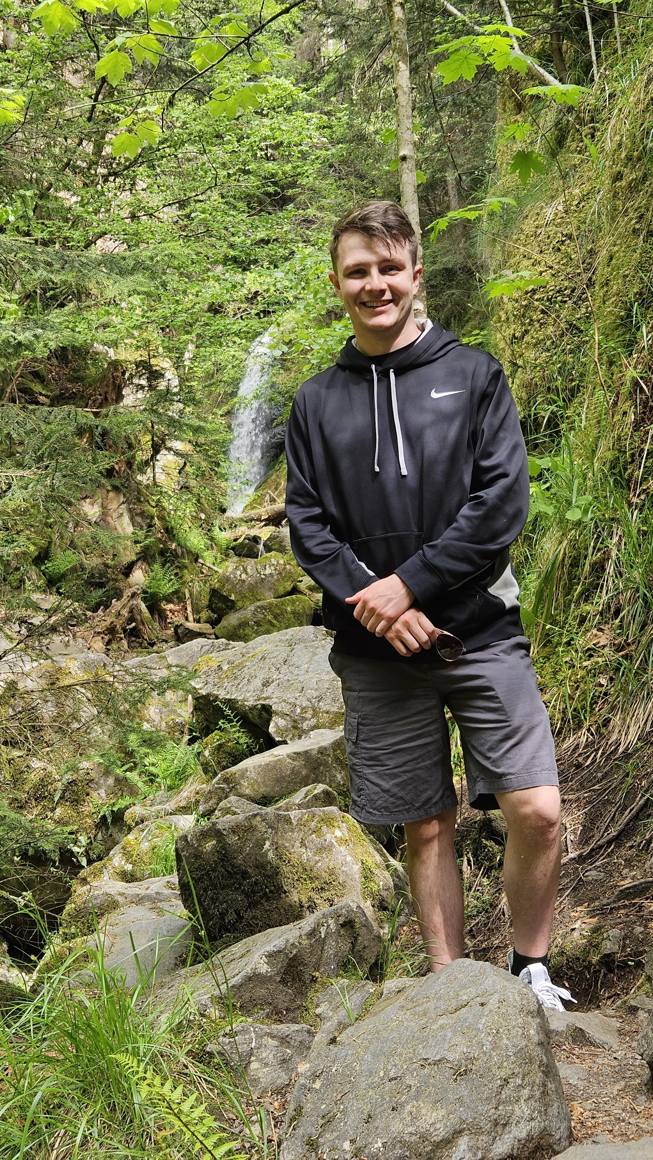 Young man with short brown hair smiling, wearing a black Nike hoodie and dark shorts, standing on rocks in a lush green forest near a small waterfall.