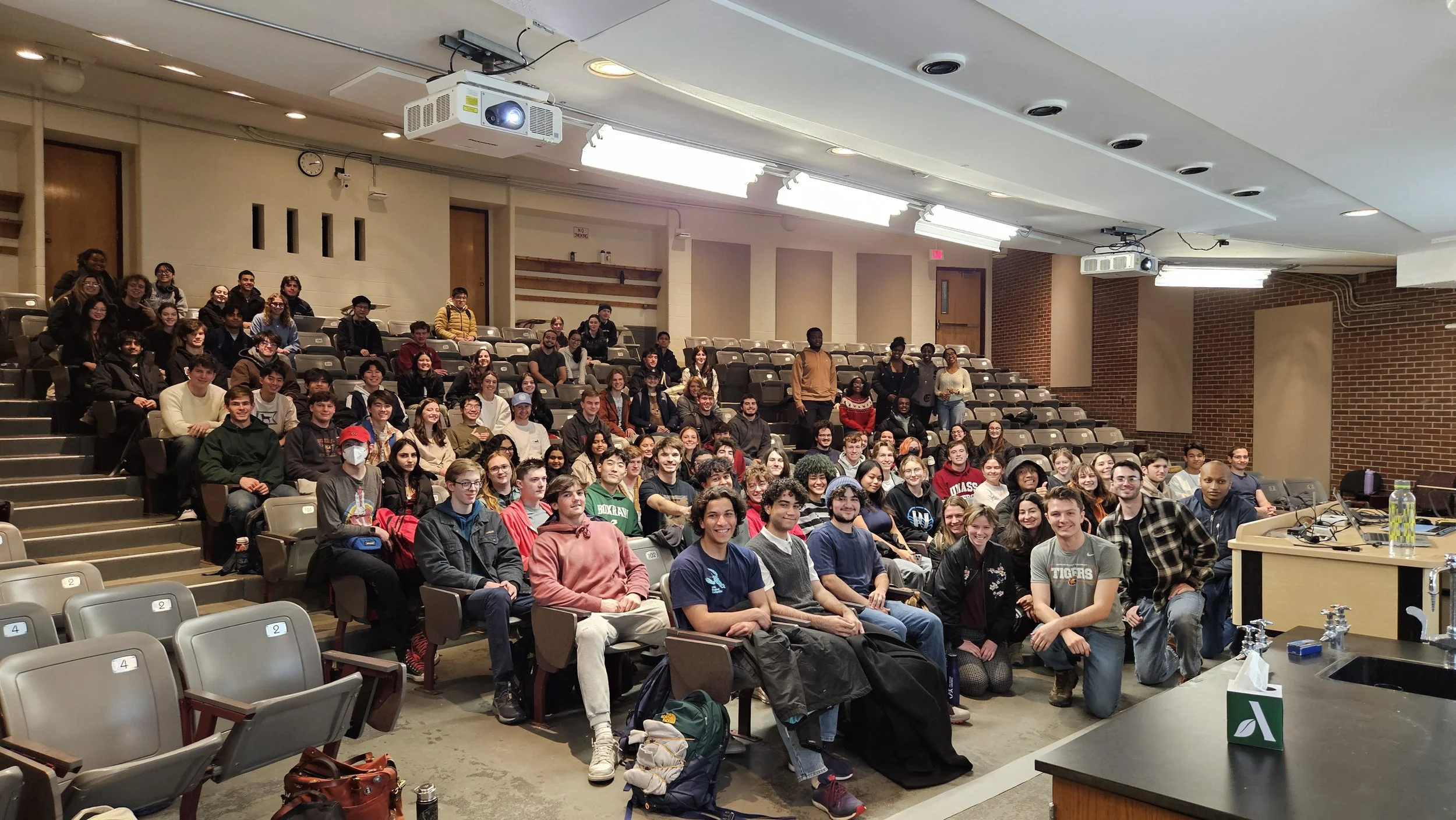 A large group of students gathered in a lecture hall, many smiling and looking at the camera. The hall has tiered seating with some empty seats, and there is laboratory equipment and water bottles on the tables in the foreground.