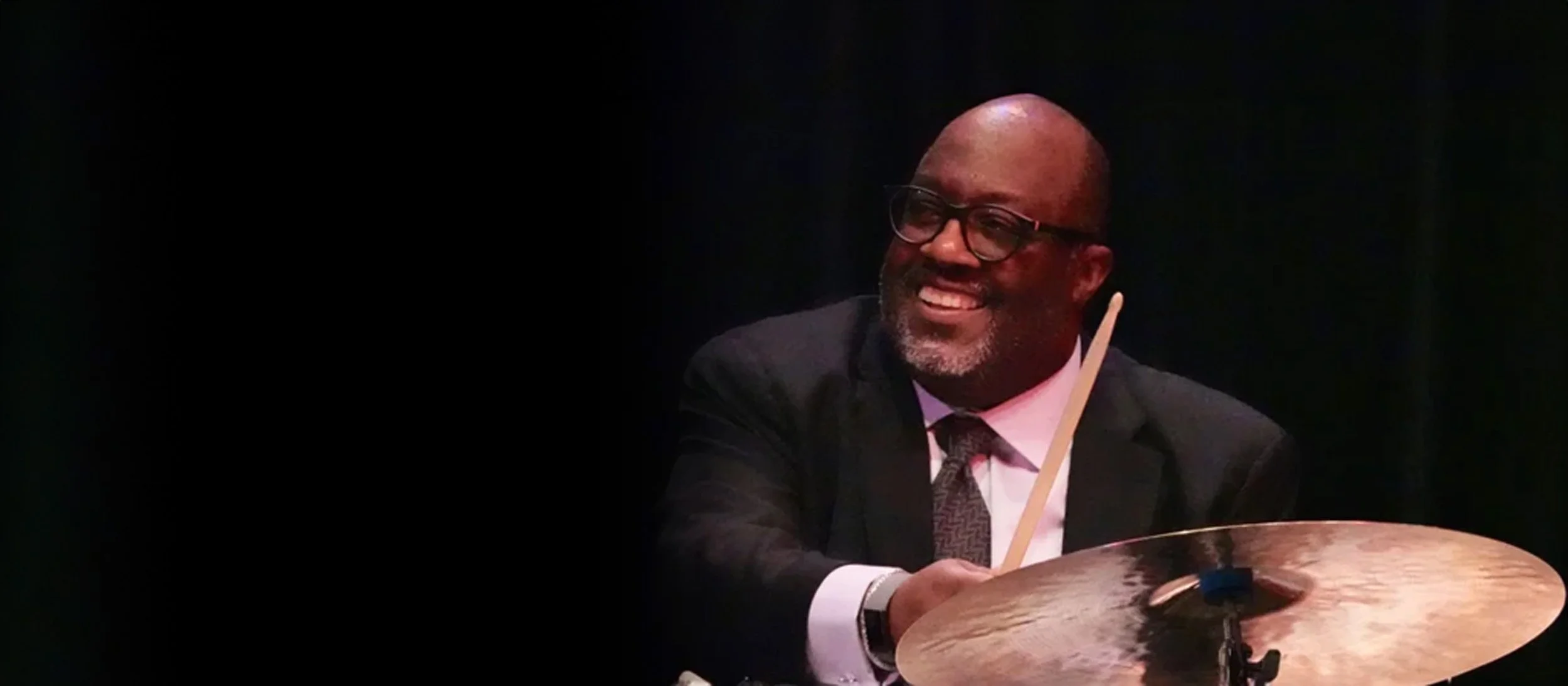 A smiling drummer wearing glasses, a dark suit, white shirt, and patterned tie, sitting at a drum set.