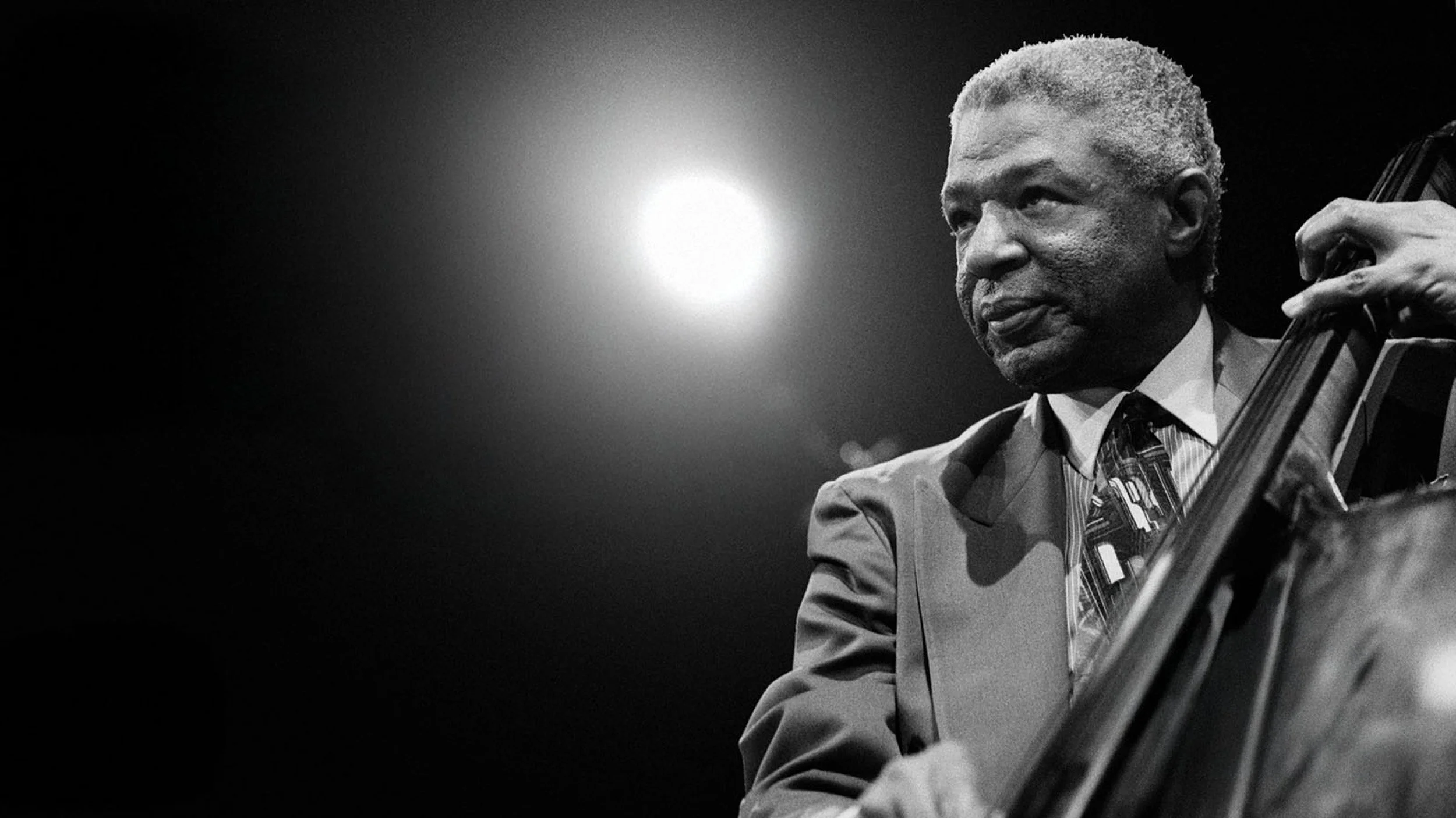 Black and white photo of an elderly man in a suit playing a double bass, focused and serious, with a bright light behind him.