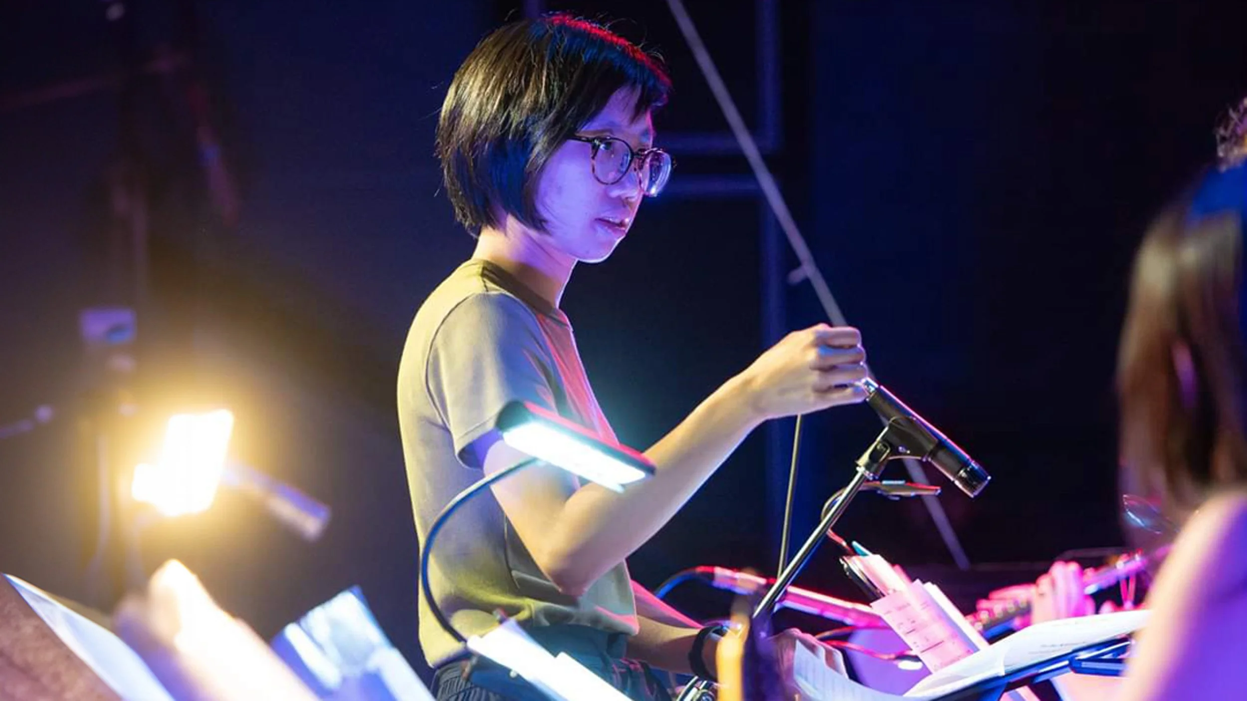 A woman conducting an orchestra on stage illuminated by colorful stage lights.
