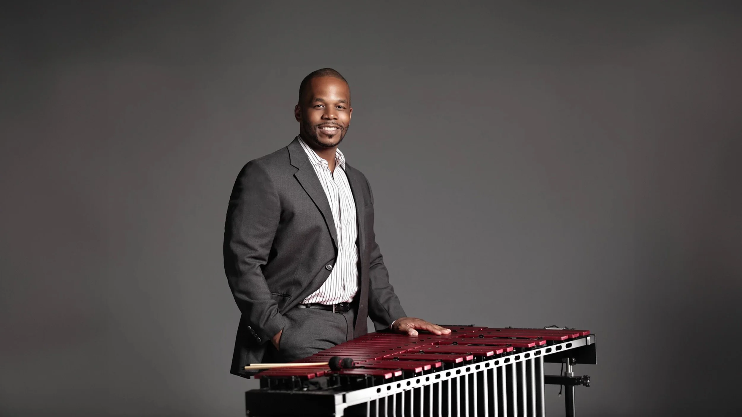 A man in a gray suit and striped shirt standing next to a red mallet and a large percussion instrument called a vibraphone, against a plain gray background.