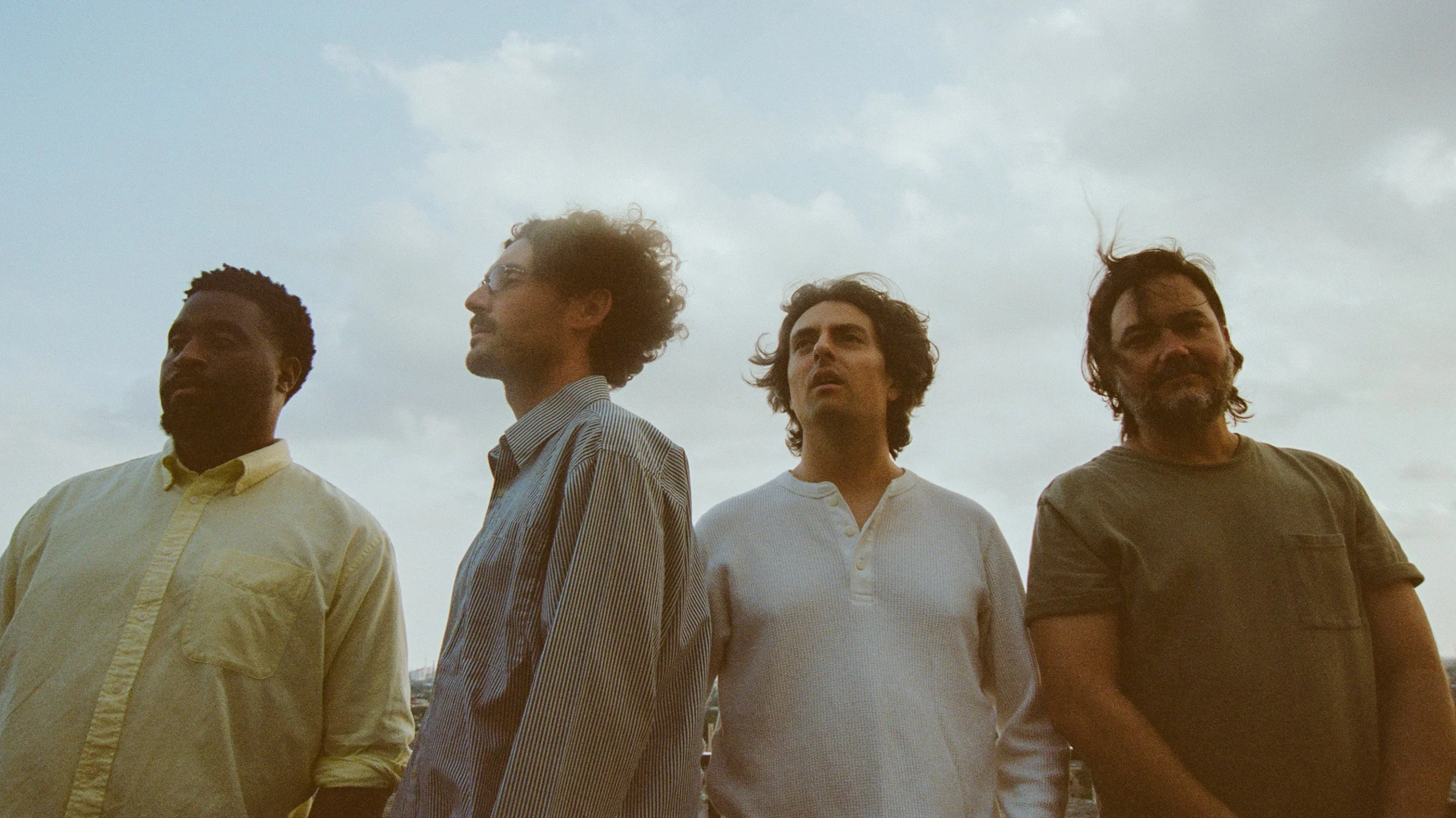 Four men standing outdoors under a cloudy sky, looking in different directions.