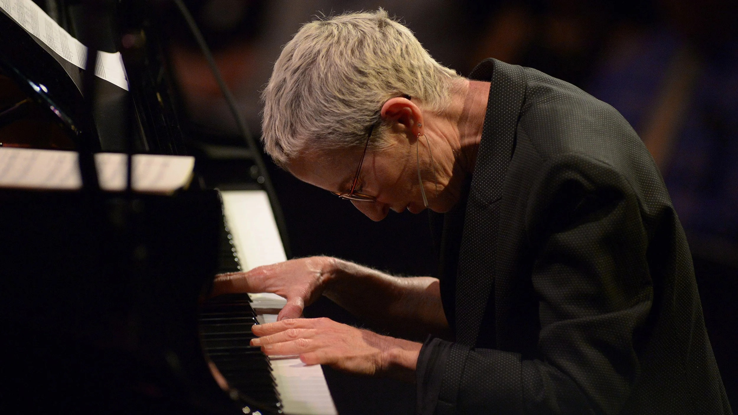 A musician with short hair and glasses playing the piano, leaning over the keyboard with a focused expression.