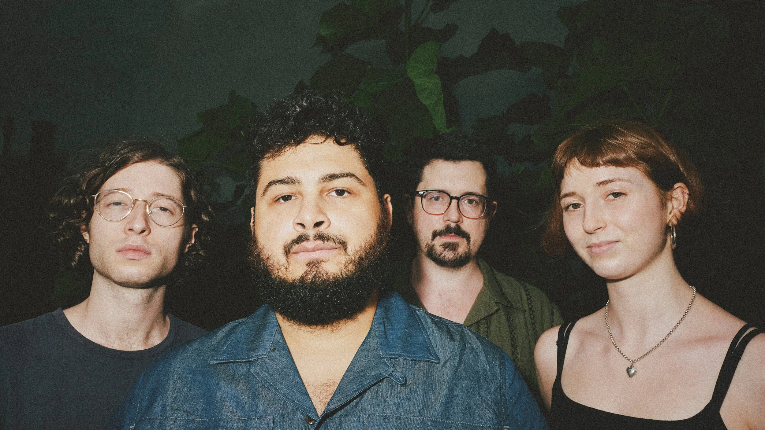 Group portrait of four people standing close together in a dark setting, with greenery in the background. Two men have glasses, one has curly hair and a beard, and one woman has short hair and wears a necklace.