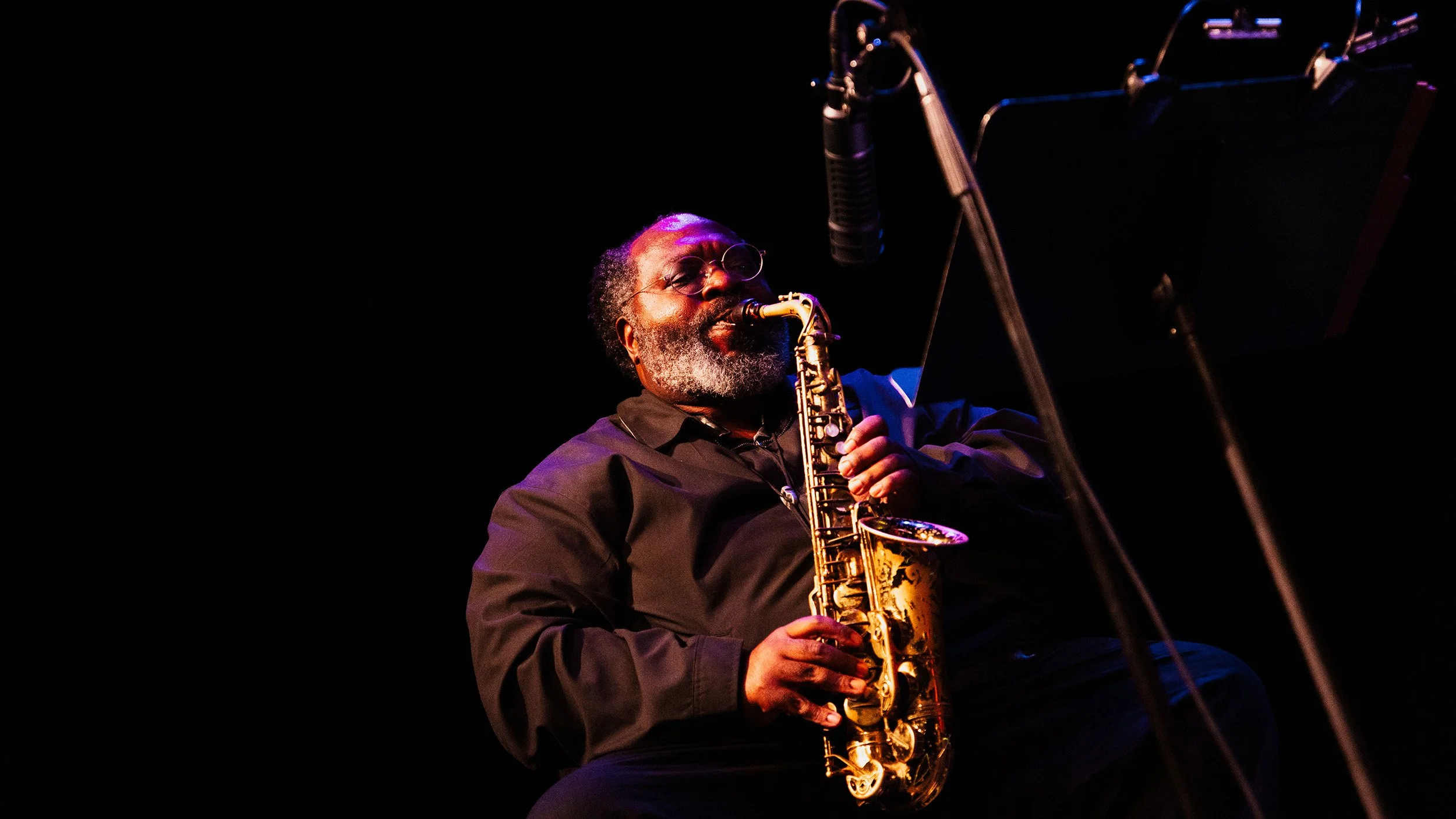 A man with glasses, a beard, and gray hair playing a gold saxophone on stage with a dark background.