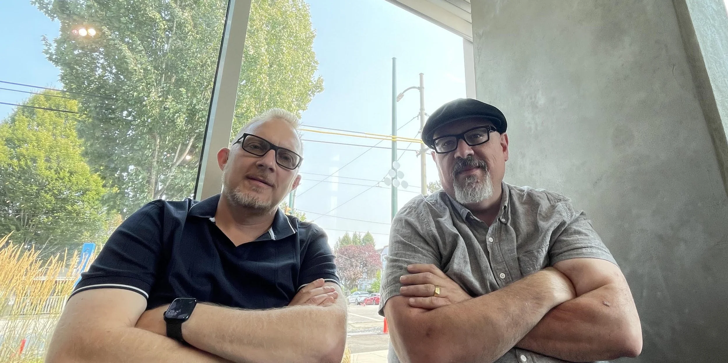 two white men wearing glasses sitting in a cafe. One is blonde, one is dark haired and wearing a hat.