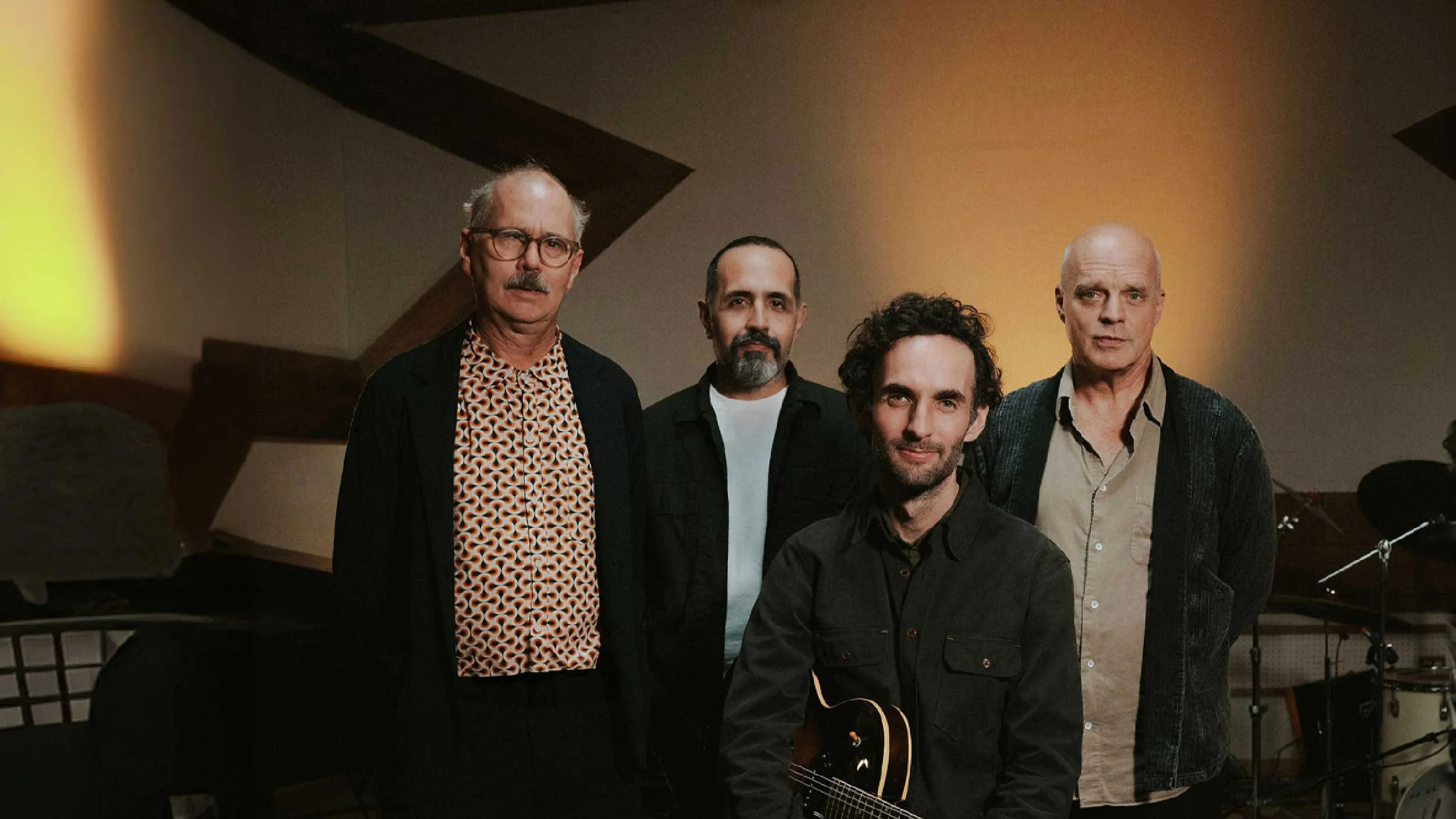 A group of five men standing indoors, with musical instruments and equipment in the background.