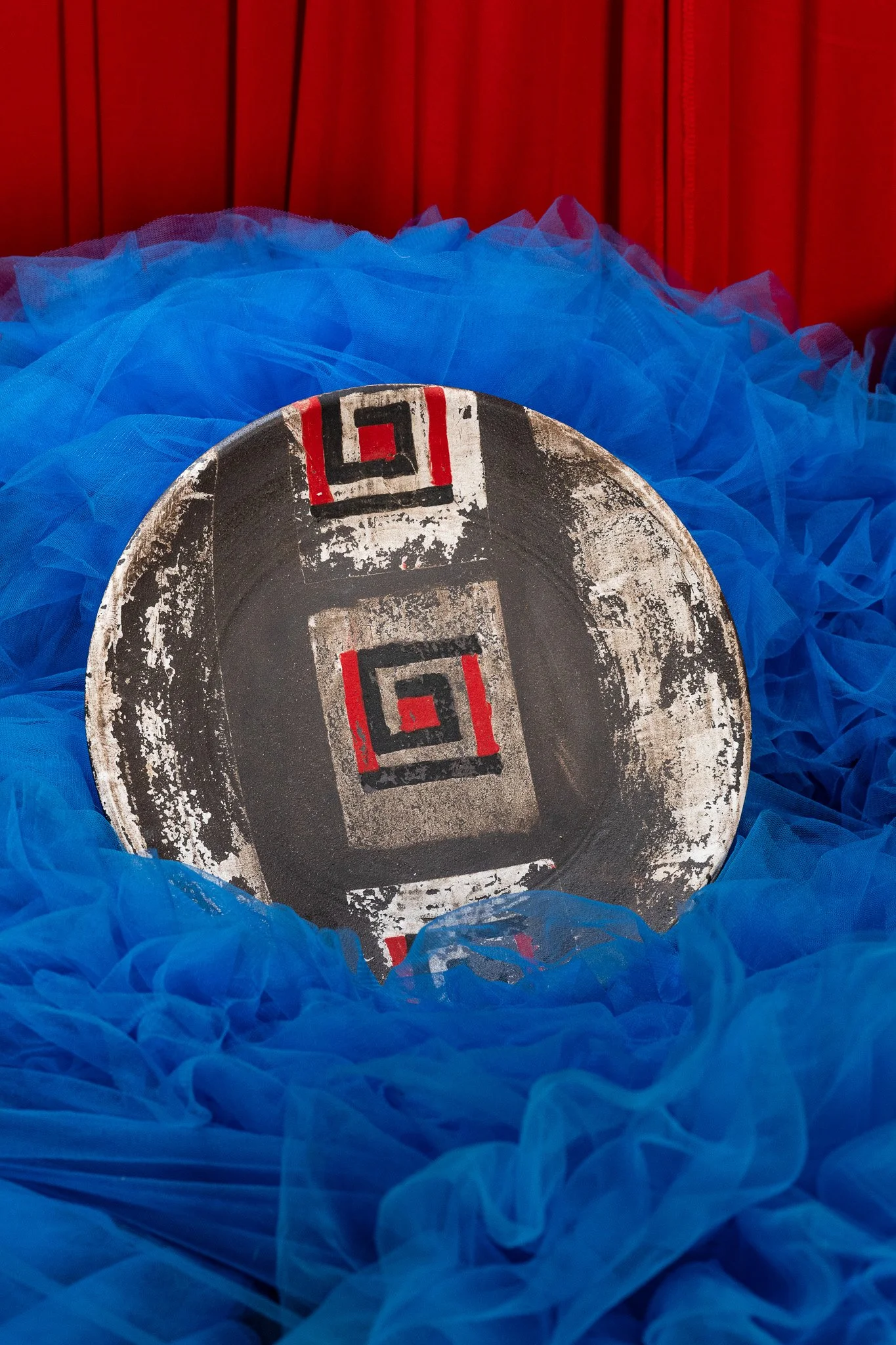 Close-up of a vintage racing wheel with a black, white, and red design, resting on a bed of bright blue tulle fabric against a red wooden background.