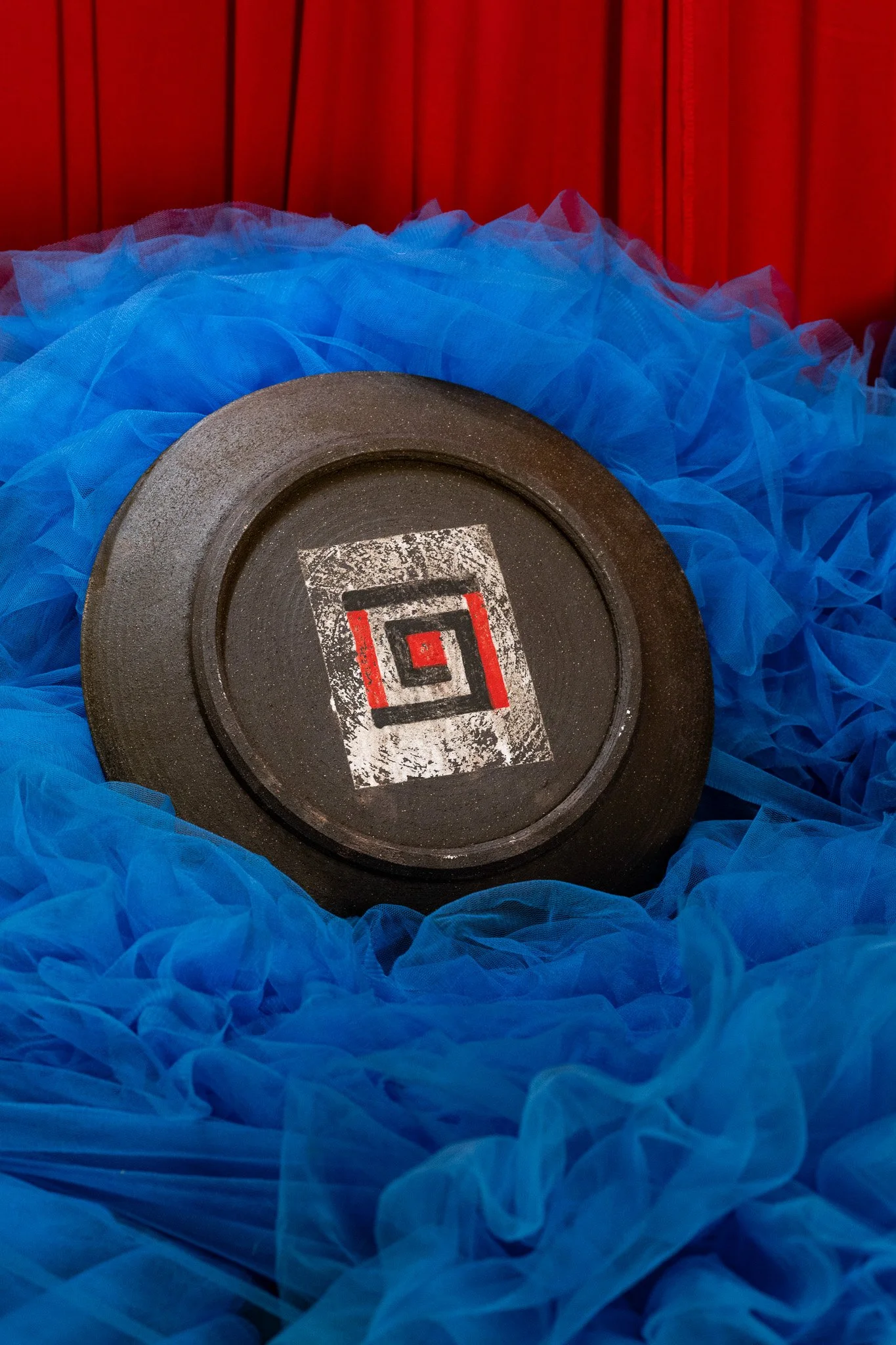 A black weight plate with a red, black, and white logo on it, resting in bright blue tulle fabric with a red curtain background.