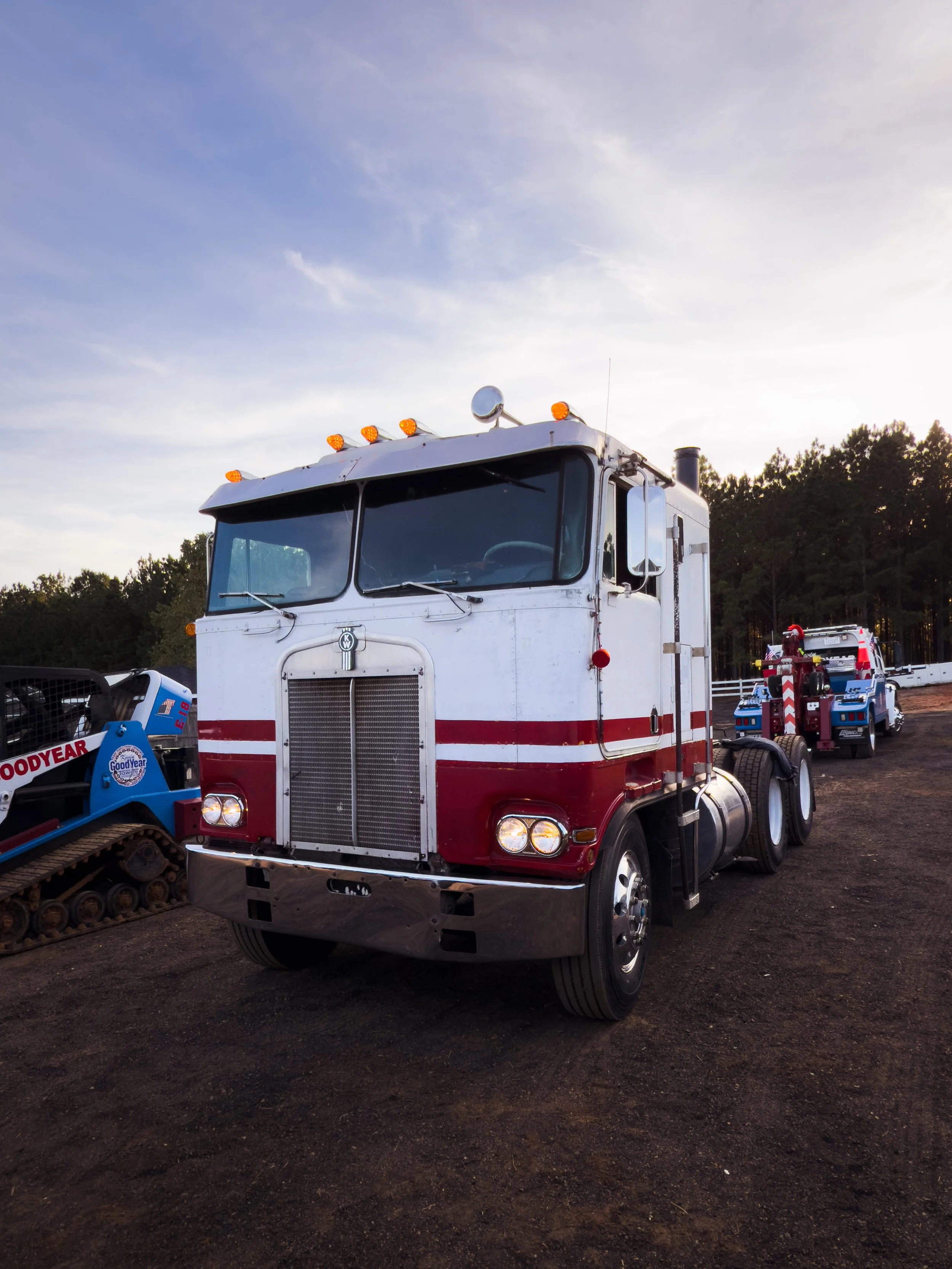 A white and red semi-truck parked on a dirt lot with other vehicles and trees in the background during sunset.
