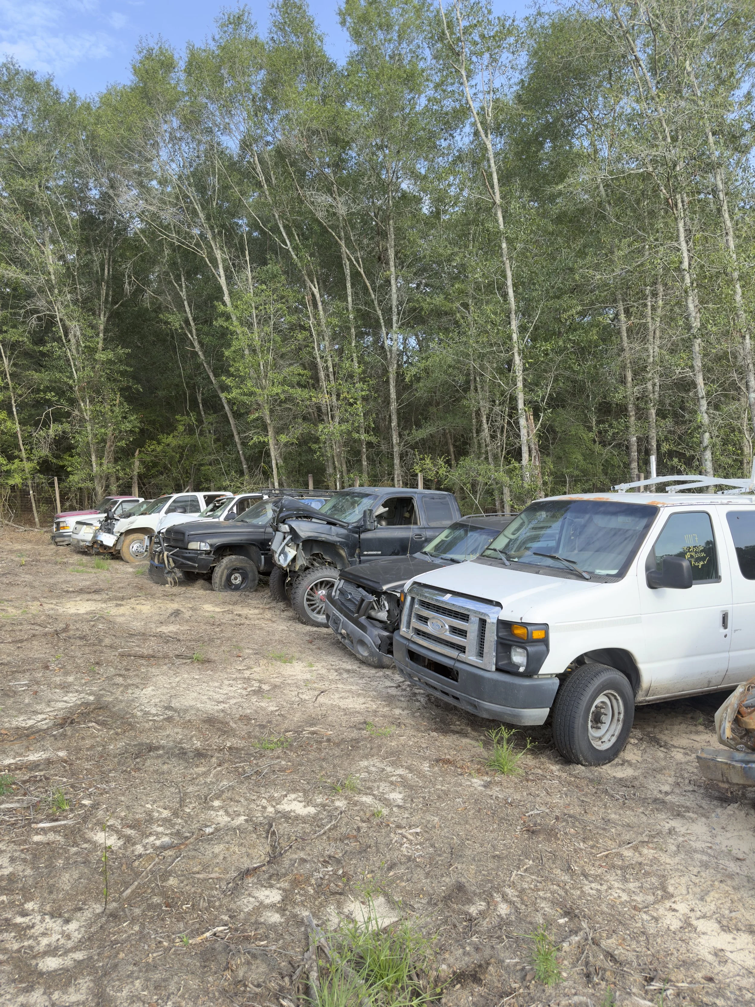 Line of damaged and wrecked vehicles, mostly pickup trucks, parked on dirt ground in front of a wooded area with green trees and a partly cloudy sky.
