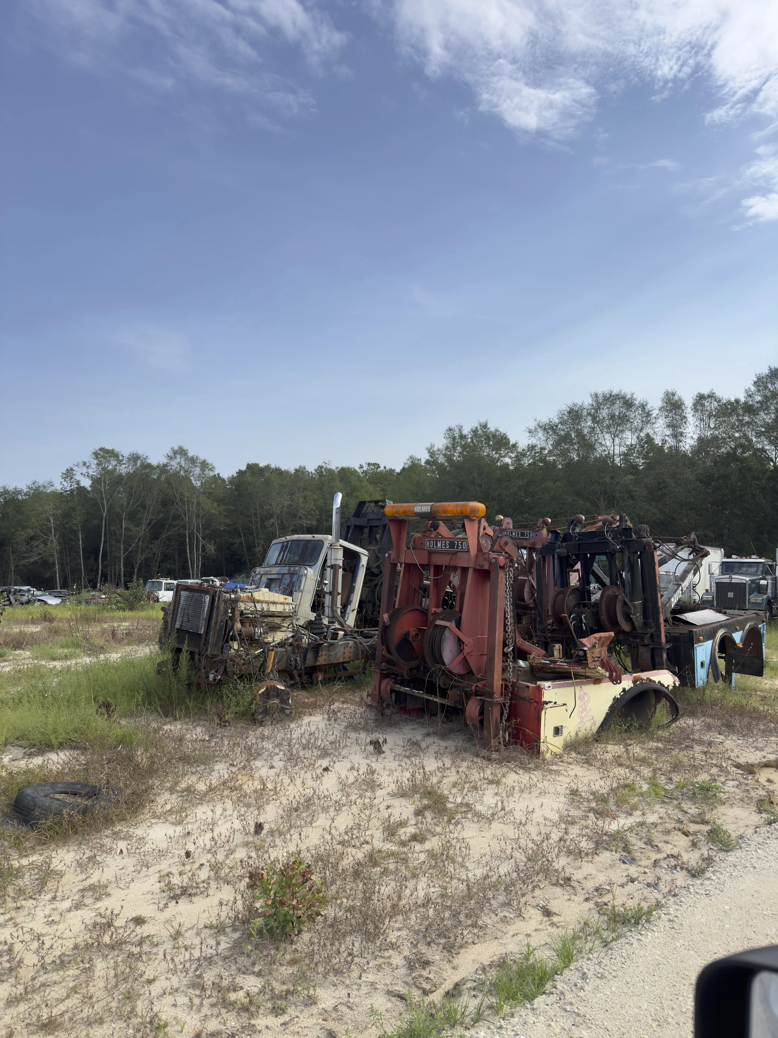 A junkyard with abandoned, rusted vehicles and equipment on a sandy, grassy field under a partly cloudy sky.