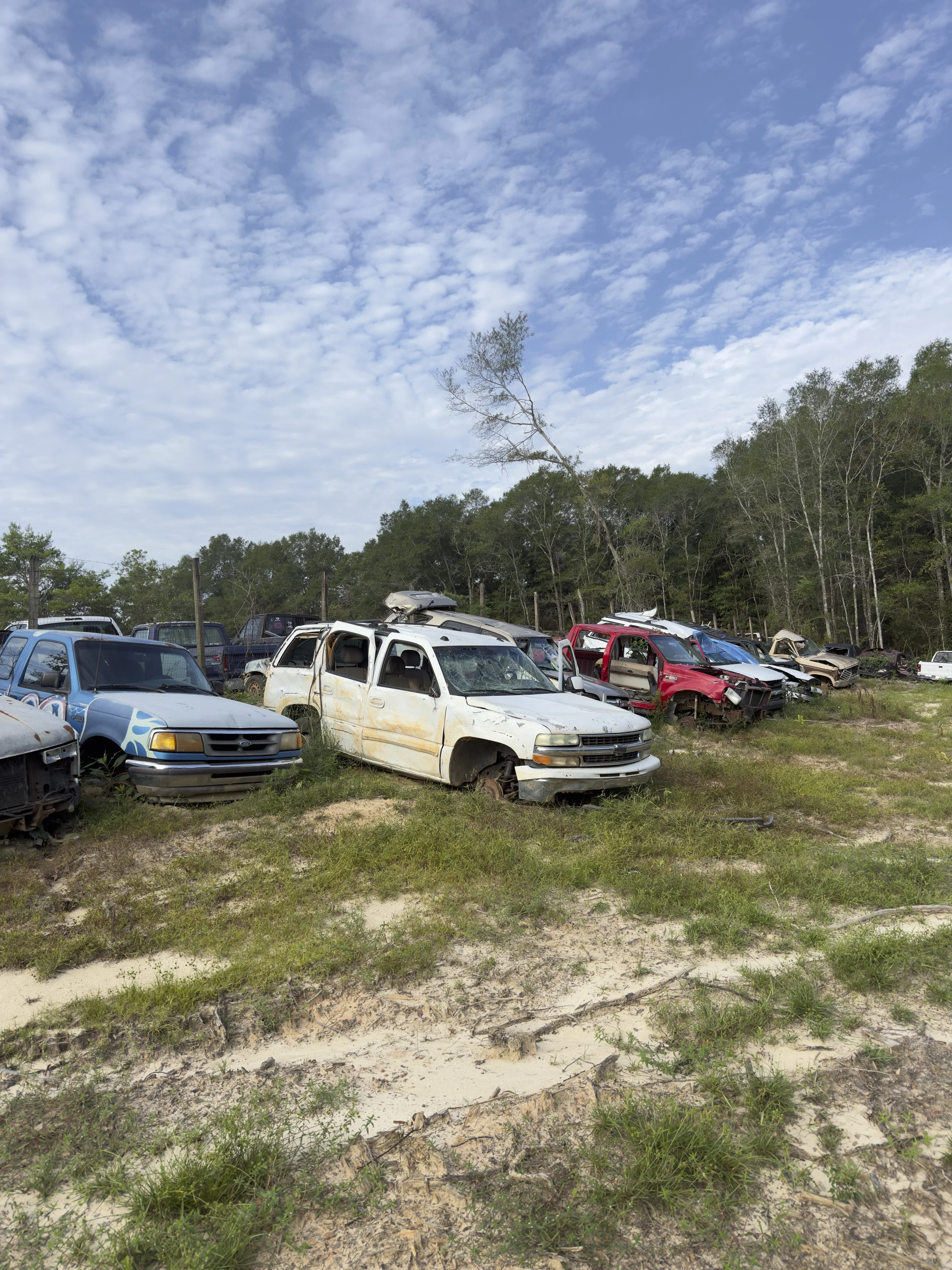 A large outdoor junkyard filled with old, damaged cars placed on grassy ground under a sky with scattered clouds and trees in the background.