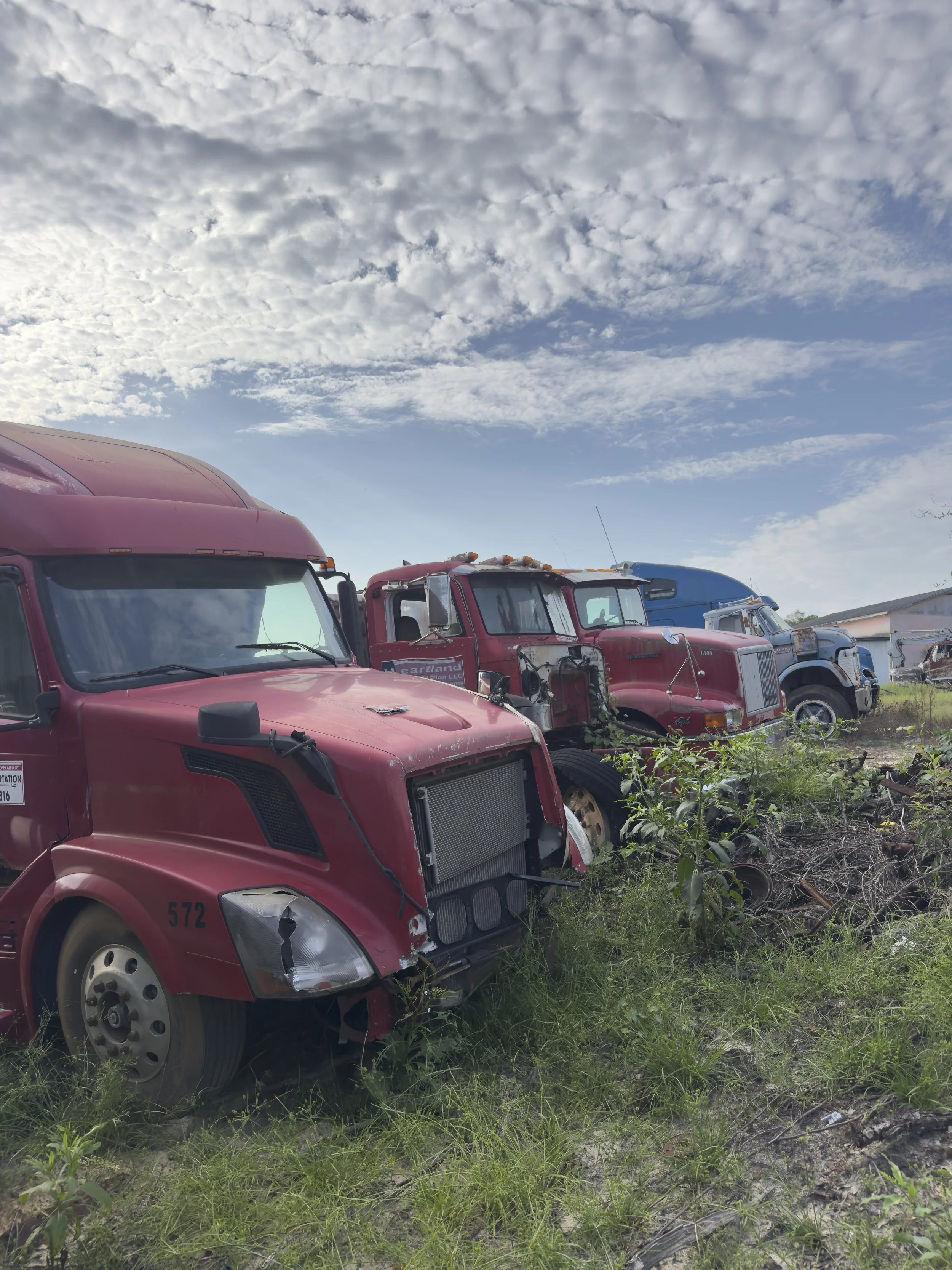 A row of old, damaged semi-trucks with various colors, parked on a grassy area under a partly cloudy sky.