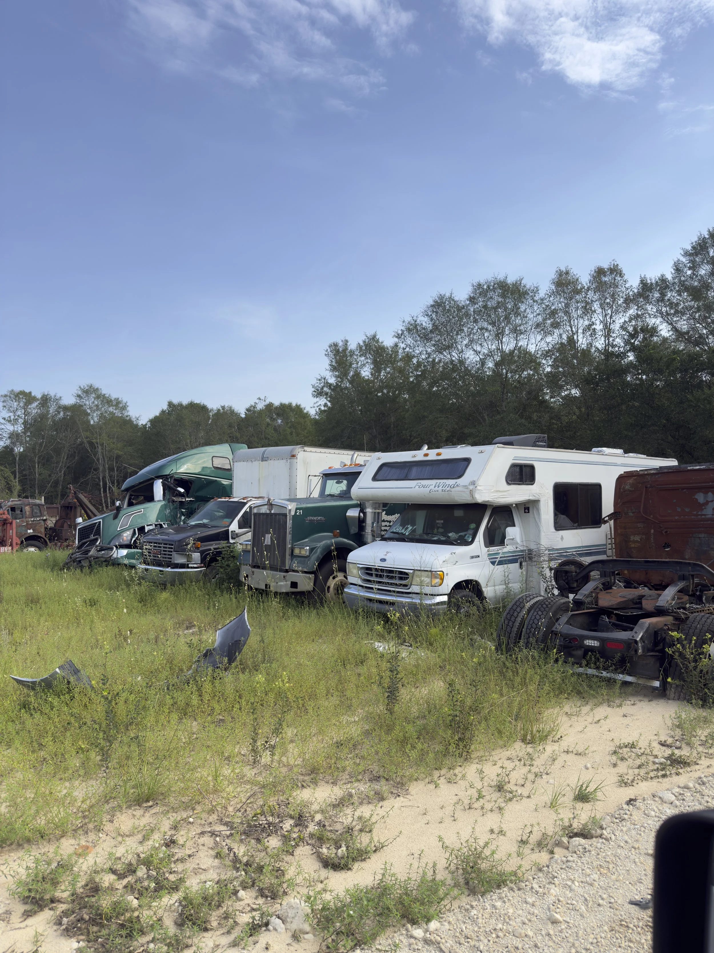 A row of various old, wrecked vehicles including trucks and a camper in a grassy, semi-rural area with trees and a blue sky overhead.