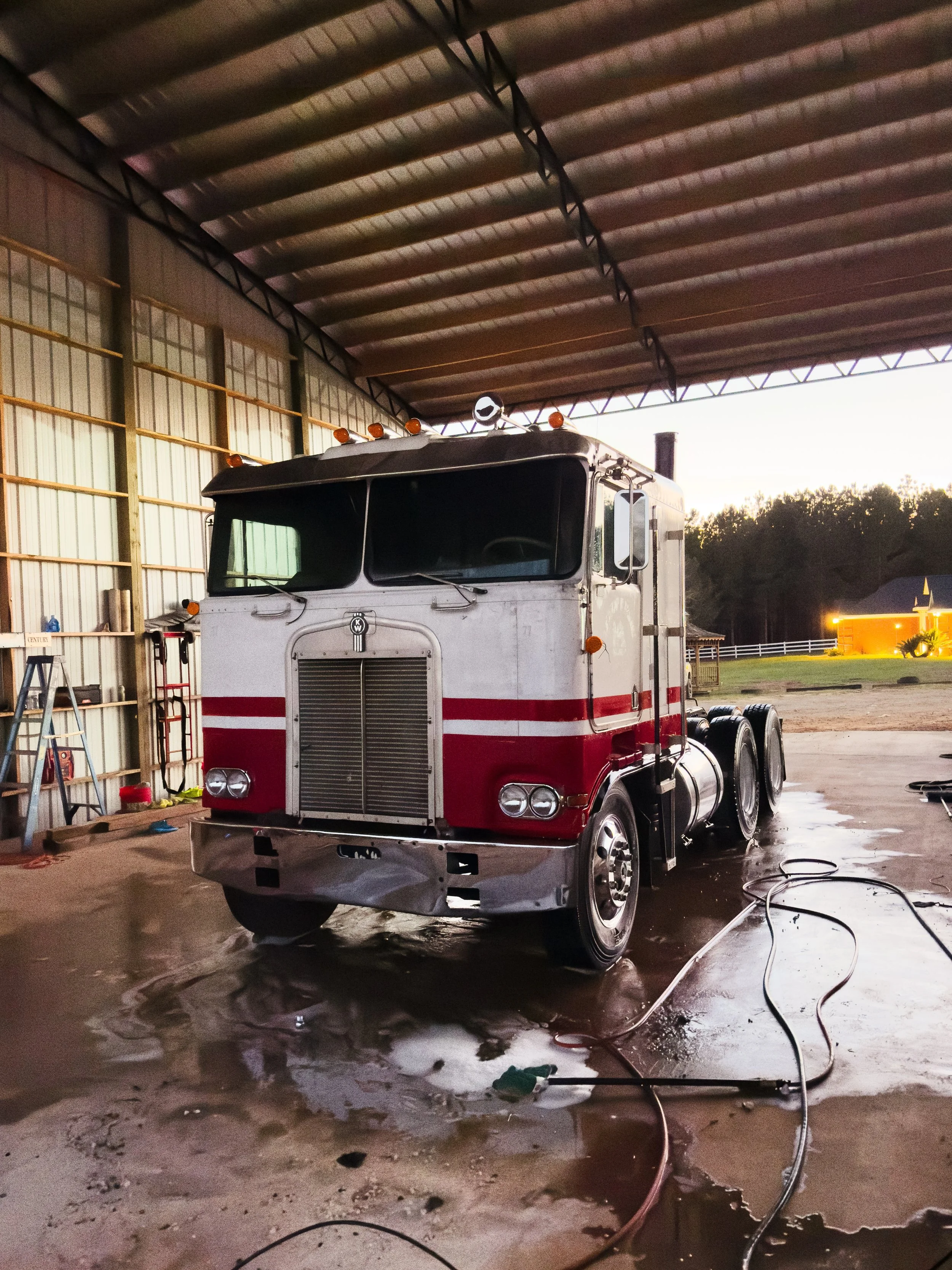 A large white and red semi-truck parked inside a metal building with a high ceiling. The truck is being washed, with water and soap on the floor, and hoses visible on the ground.