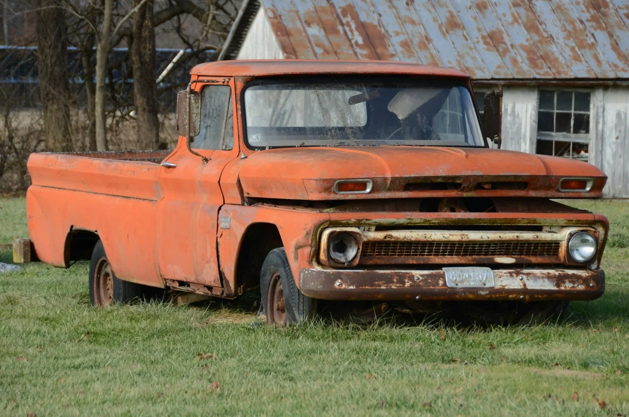 Old, rusty orange pickup truck parked on grass in front of a weathered wooden building with a rusted metal roof and leafless trees in the background.
