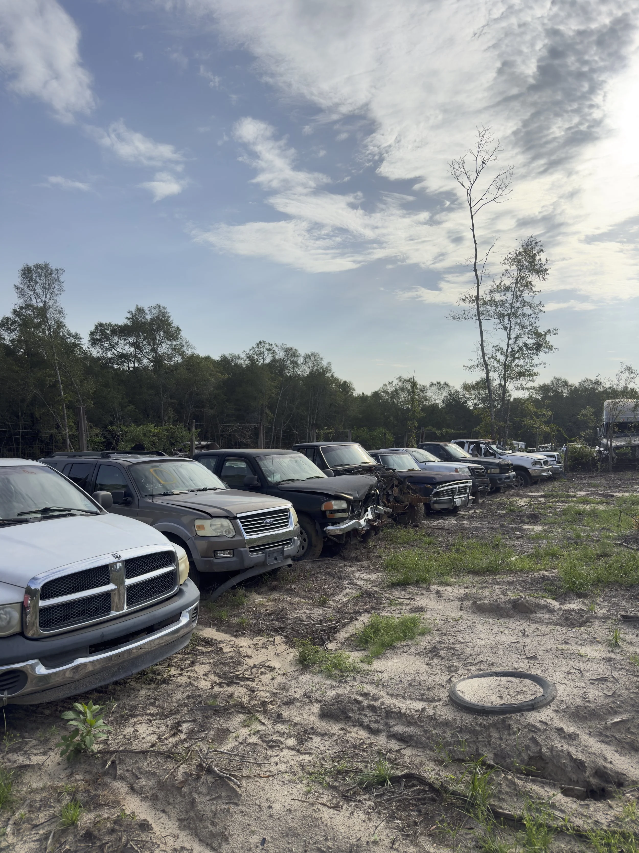 A line of old, damaged, and abandoned vehicles parked on a dirt lot in front of a wooded area. The sky is partly cloudy.