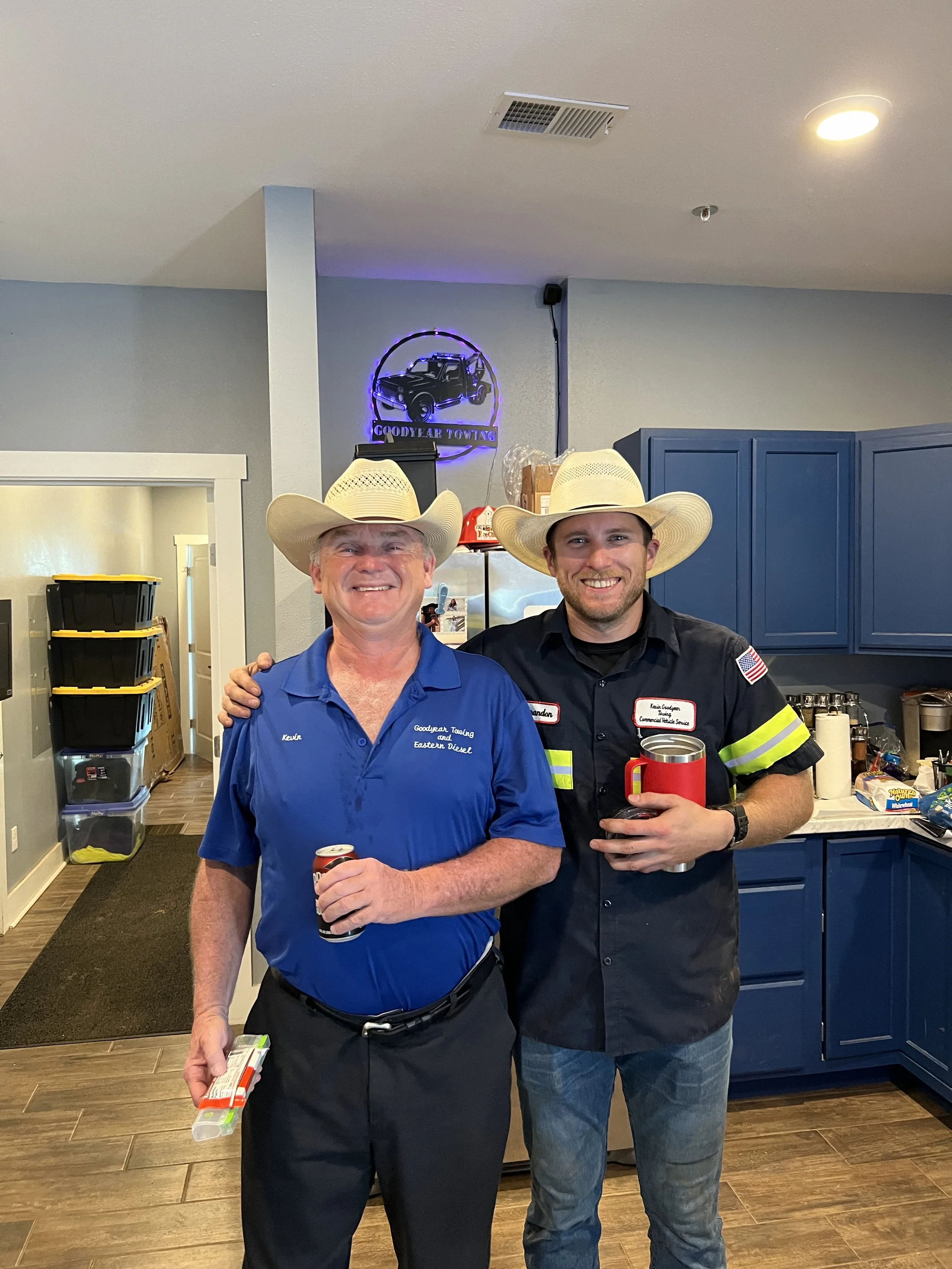 Two smiling men wearing cowboy hats, one in a blue shirt and the other in a black uniform with a name tag and American flag patch, standing indoors in front of blue kitchen cabinets. They are holding cans of soda and have their arms around each other's shoulders.