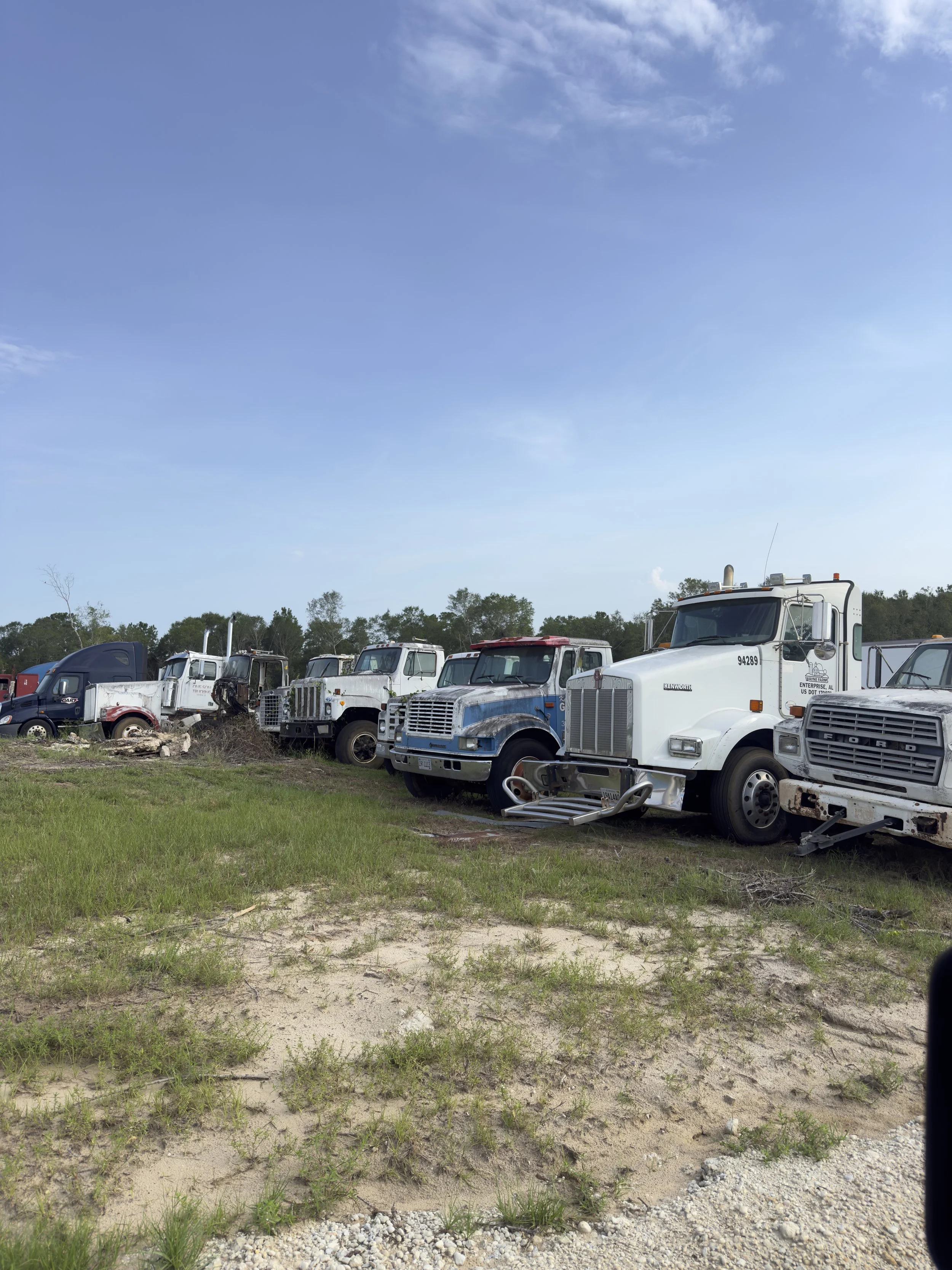 A row of old, damaged semi-trucks and vehicles parked on a grassy and sandy lot under a partly cloudy sky.