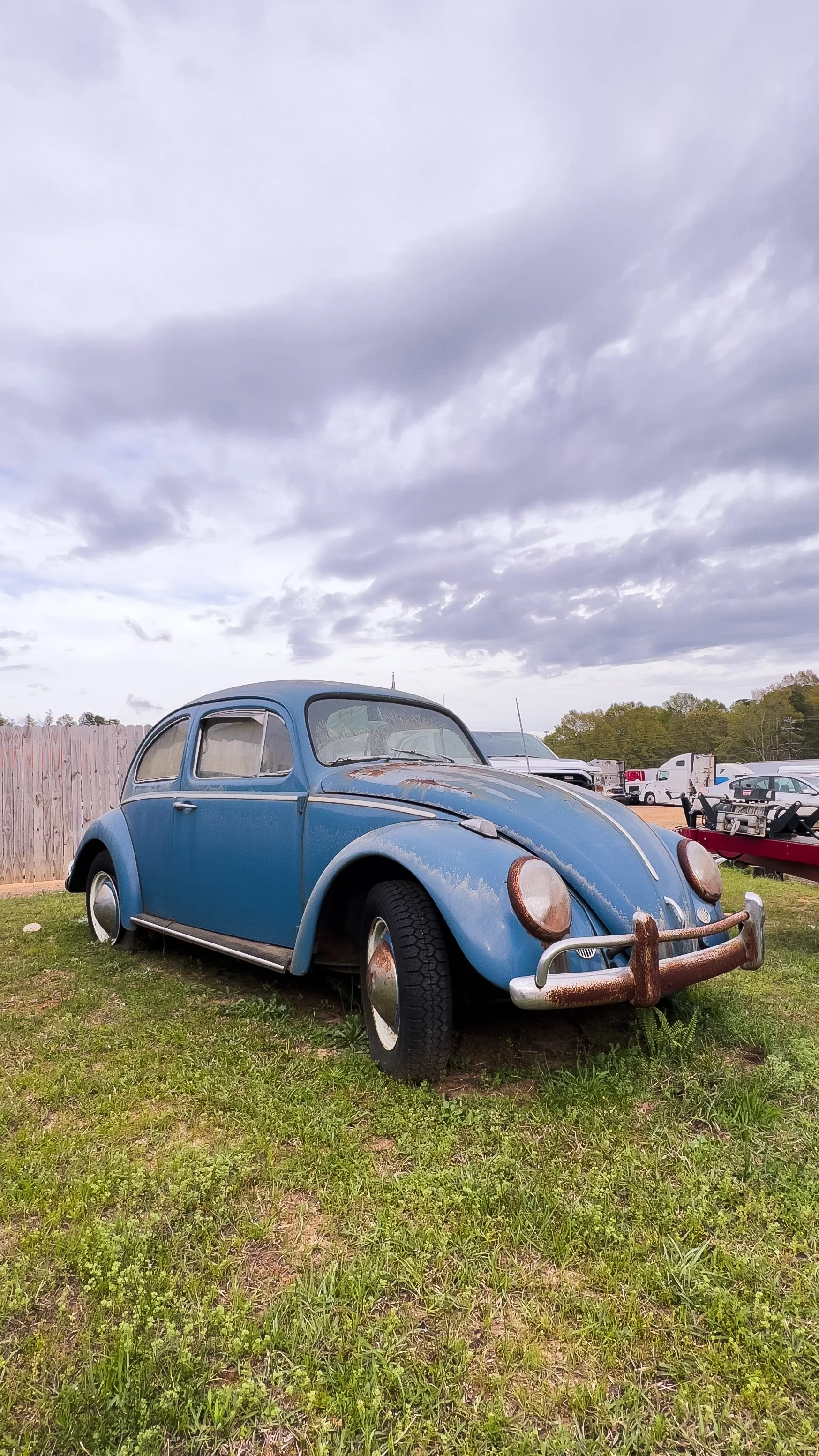 An old, blue Volkswagen Beetle car parked on grass before a wooden fence and a cloudy sky, with other vehicles and trailers in the background.