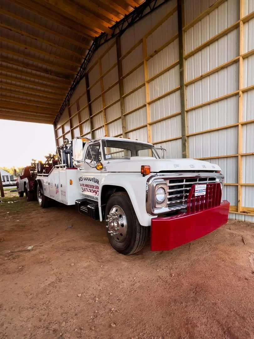 A white tow truck parked inside a large wooden building with a curved metal roof, with a red bumper guard attached to the front.