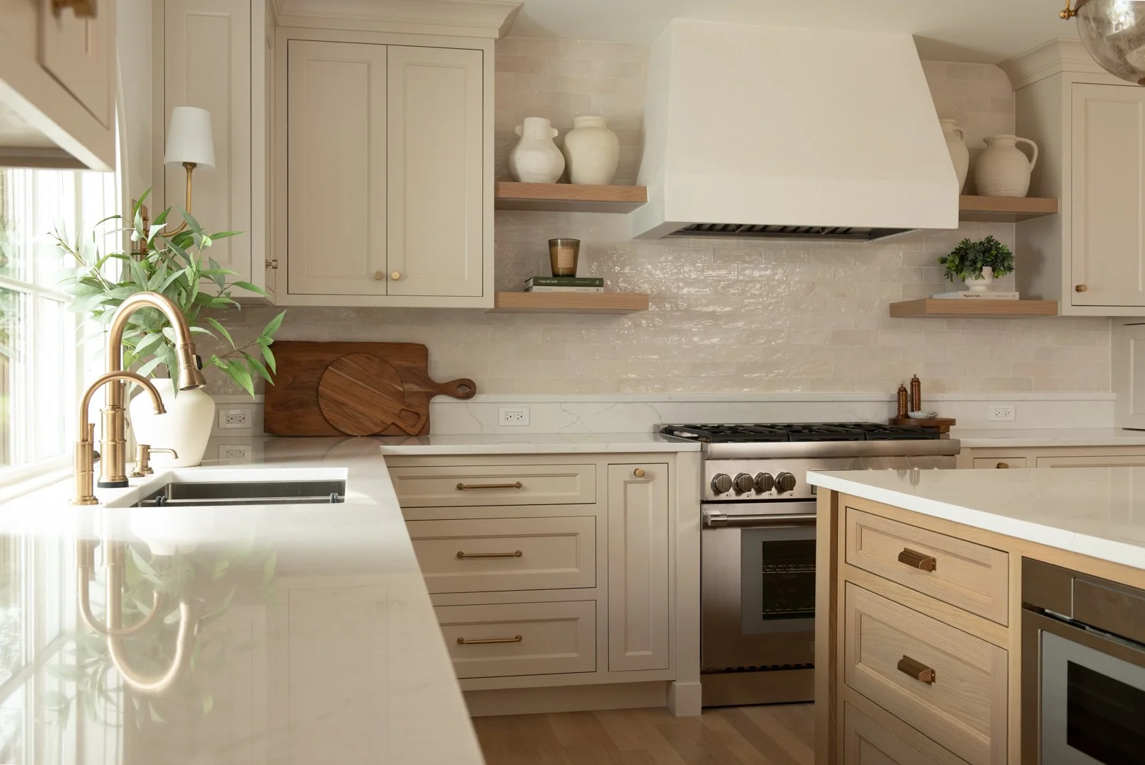 Modern kitchen with white cabinetry, gold fixtures, beige backsplash, and wooden accents. Features a window with natural light, a sink, and a stove with an overhead vent hood.