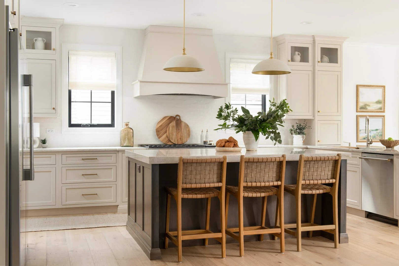 Bright kitchen with white cabinets, marble countertops, a kitchen island with wooden bar stools, plants, and natural light from windows.