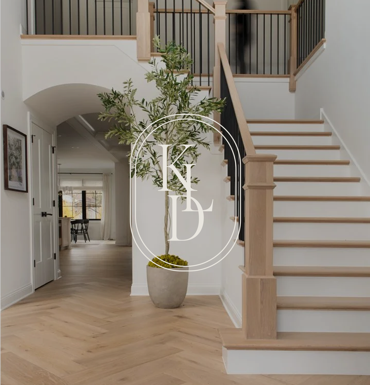 Interior of a house with a staircase, potted indoor plant, and hallway leading to a dining area with window.
