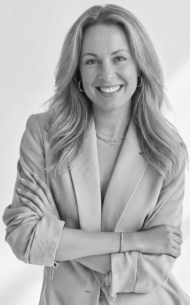 Black and white portrait of a smiling woman with wavy hair, wearing a blazer, layered necklaces, hoop earrings, and a bracelet, crossing her arms.