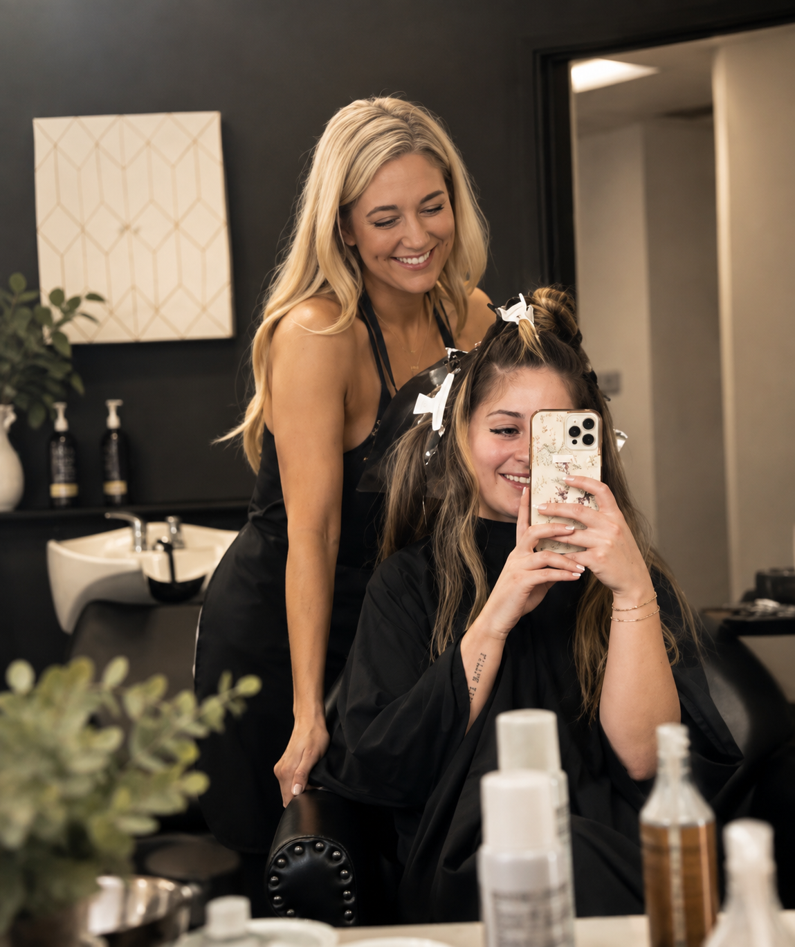A woman with long blonde hair and a black apron is styling another woman's hair with curlers in a salon. The woman getting her hair styled is sitting in a chair, holding a smartphone and smiling, with a salon cape on. The background shows salon sinks, bottles, and modern decor.