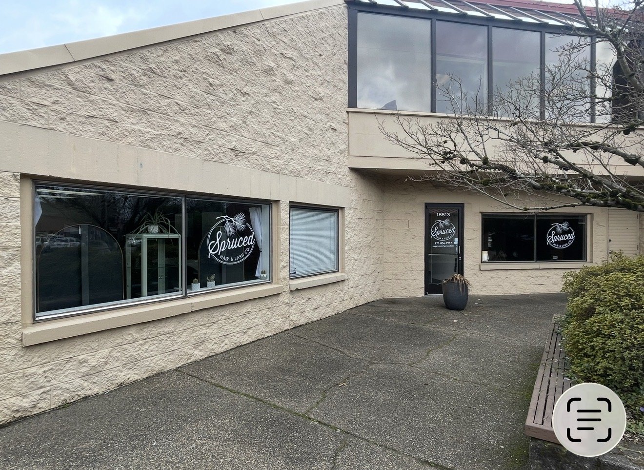 Outside view of a hair salon named Sprucer, with a beige brick exterior. The salon has two large windows displaying the Sprucer logo, and a glass door with the salon's name and phone number. A potted plant is near the entrance, and there is a bench and shrubbery to the right.