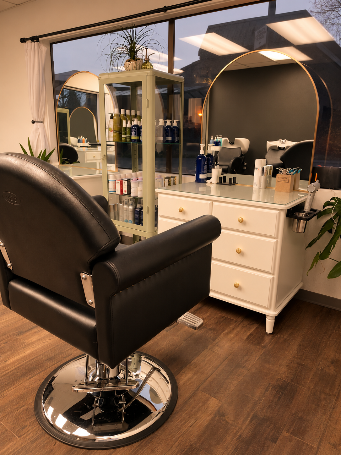 A modern salon station with a black salon chair facing a white cabinet with drawers, topped with bottles of hair products, a mirror, and various hair styling tools, with large windows in the background.