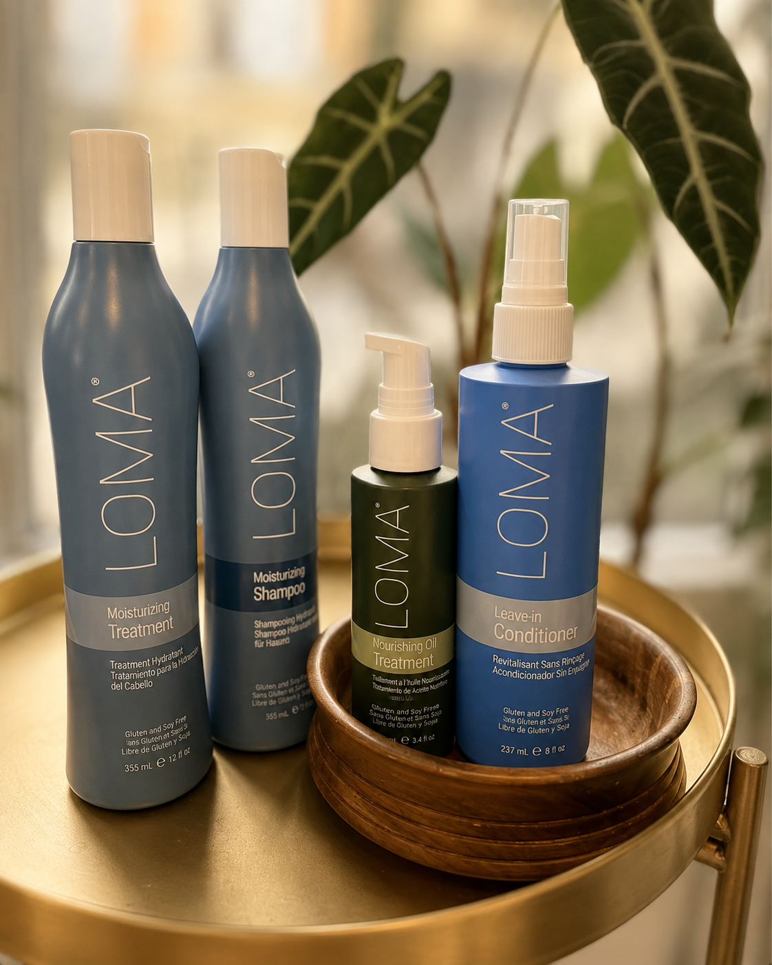 Four LO:MÁ hair care bottles on a gold round table with a wooden bowl. Two are labeled moisturizing treatment and sulfate-free shampoo, one nourishing oil treatment, and one leave-in conditioner. A leafy plant is in the background.