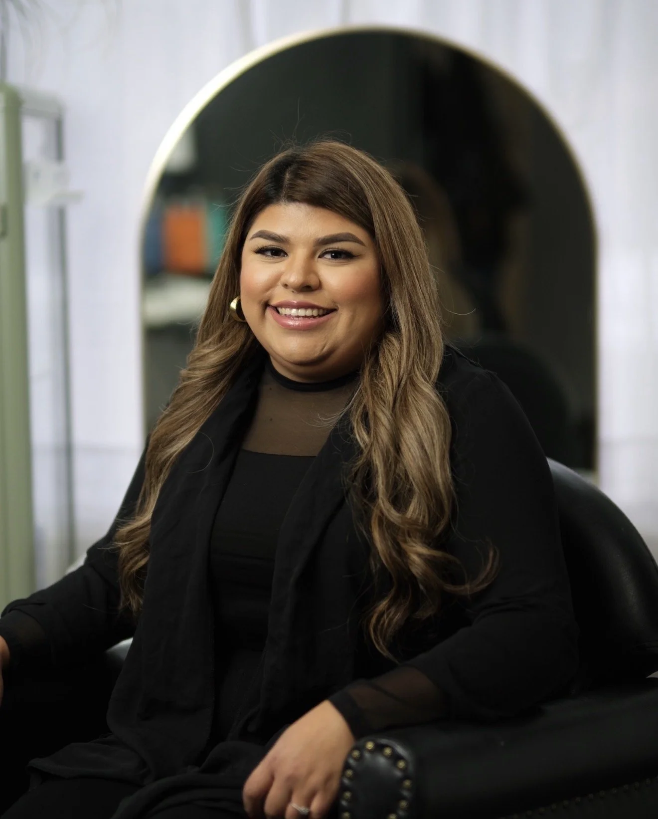 A woman with long brown wavy hair, wearing a black blazer and a sheer black top, smiling at the camera, sitting in a black chair in a modern indoor setting.