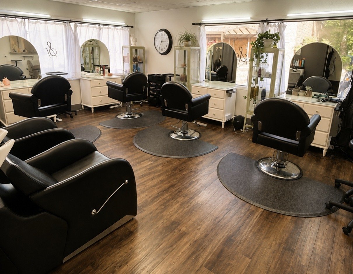 Empty hair salon with black chairs, white cabinets, mirrors, and wooden flooring.