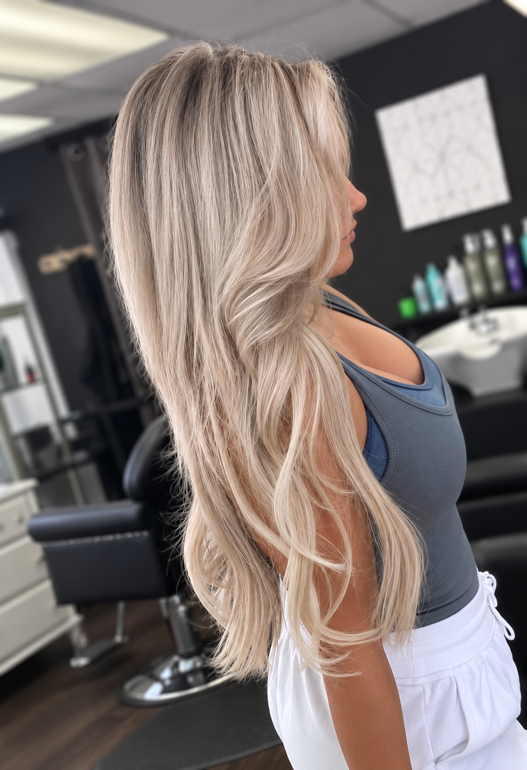 Side view of a woman with long, wavy blonde hair, wearing a blue tank top and white shorts, inside a hair salon.