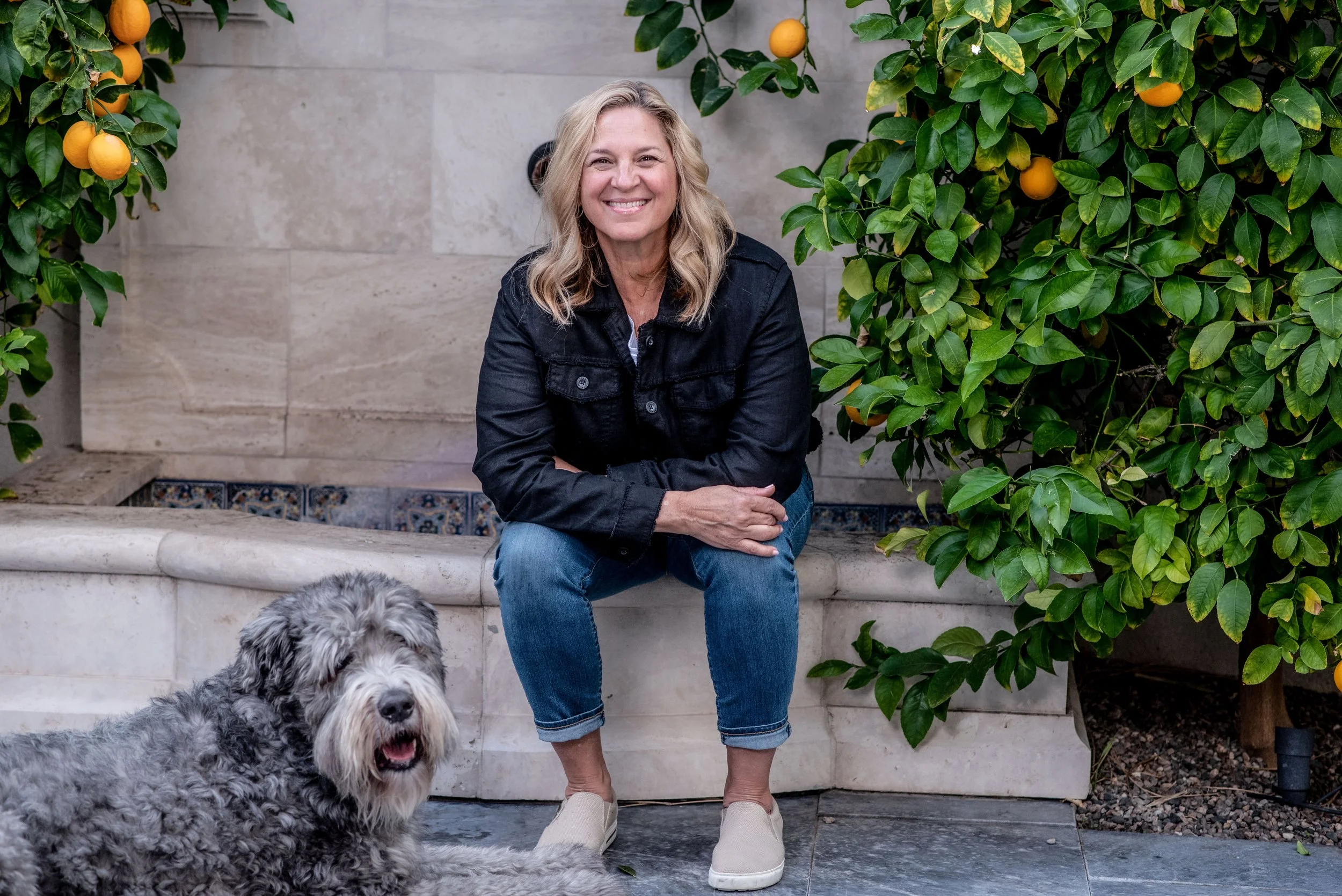 A smiling woman with blonde hair, wearing a black jacket and blue jeans, sits on a stone bench surrounded by green shrubbery with orange fruits. A large, gray, fluffy dog with a happy expression sits in front of her.