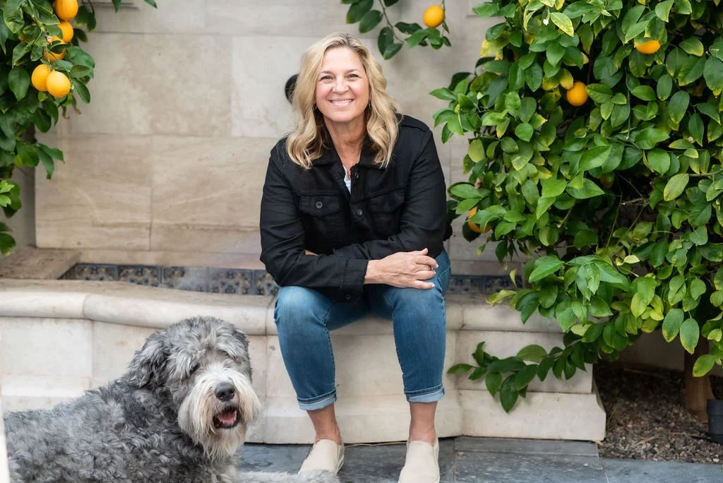 A smiling woman sitting on a stone bench next to a large, gray, curly-haired dog, with lemon trees and green foliage in the background.