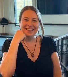 A woman with blonde hair smiling while sitting at a table indoors.