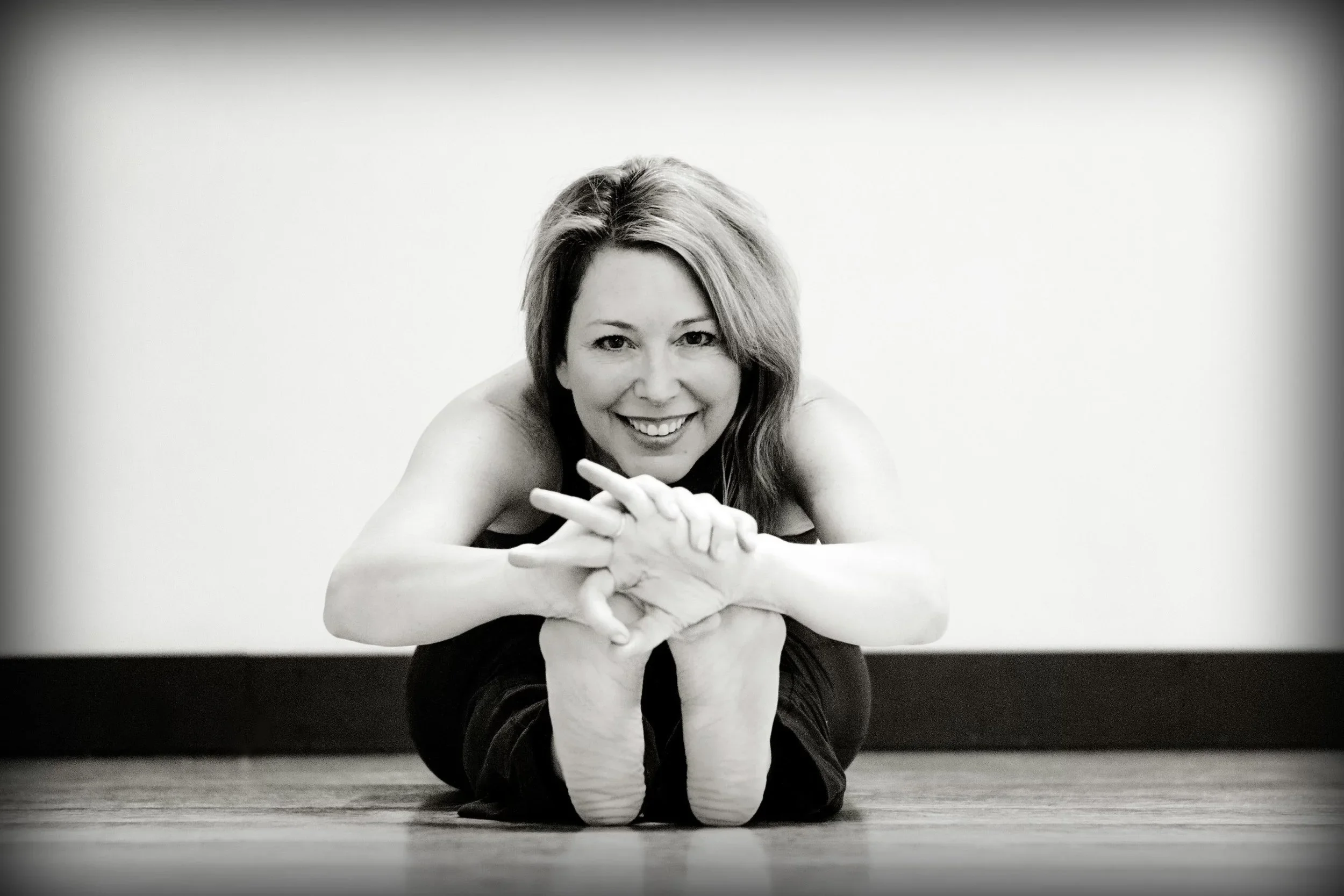 A smiling woman practicing yoga on a wooden floor, lying on her stomach with her head lifted and hands clasped in front.